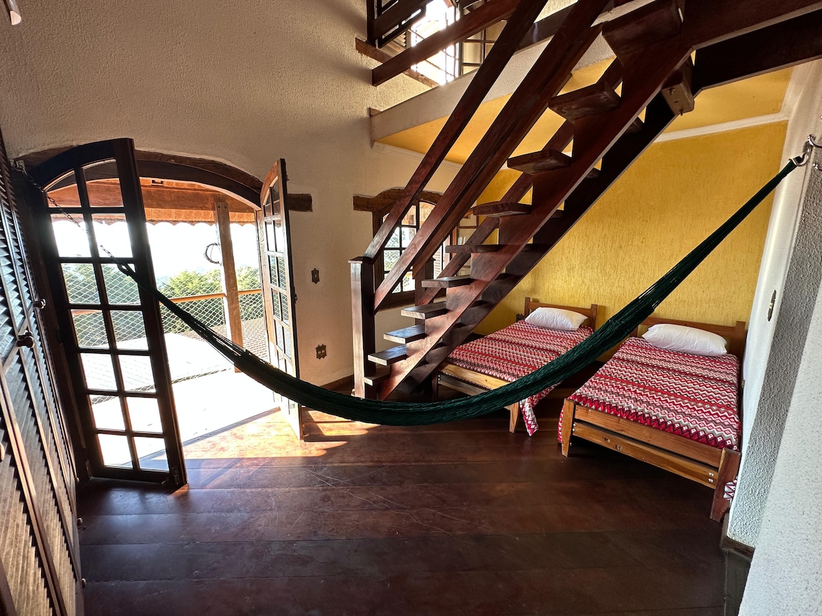 A cozy sleeping area is displayed, featuring two wooden beds with light bedding. An inviting hammock is suspended between two points, adding a relaxed touch. A set of double doors leads to an outdoor balcony, offering scenic views and natural light through the open access.