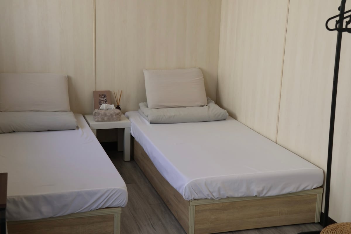 The room features two spacious beds, each with simple wooden frames and neutral-colored bedding. A small bedside table is positioned between the beds, complemented by decorative items. The light-colored walls contribute to a bright and comfortable environment.