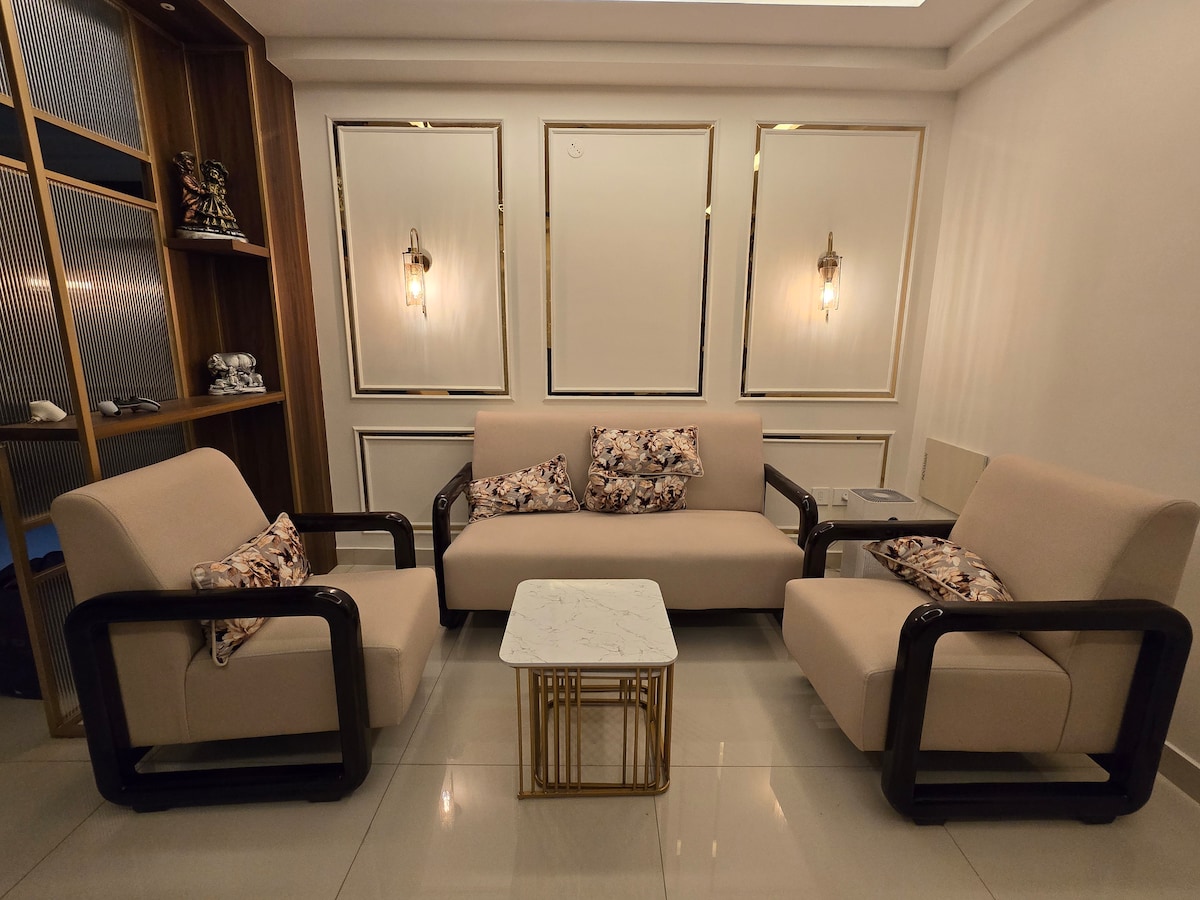 A stylish living area is showcased with neutral-colored sofas and matching armchairs, complemented by decorative cushions. A simple white marble coffee table is positioned at the center, while the walls feature elegant light fixtures and mirrored accents, enhancing the modern interior design.