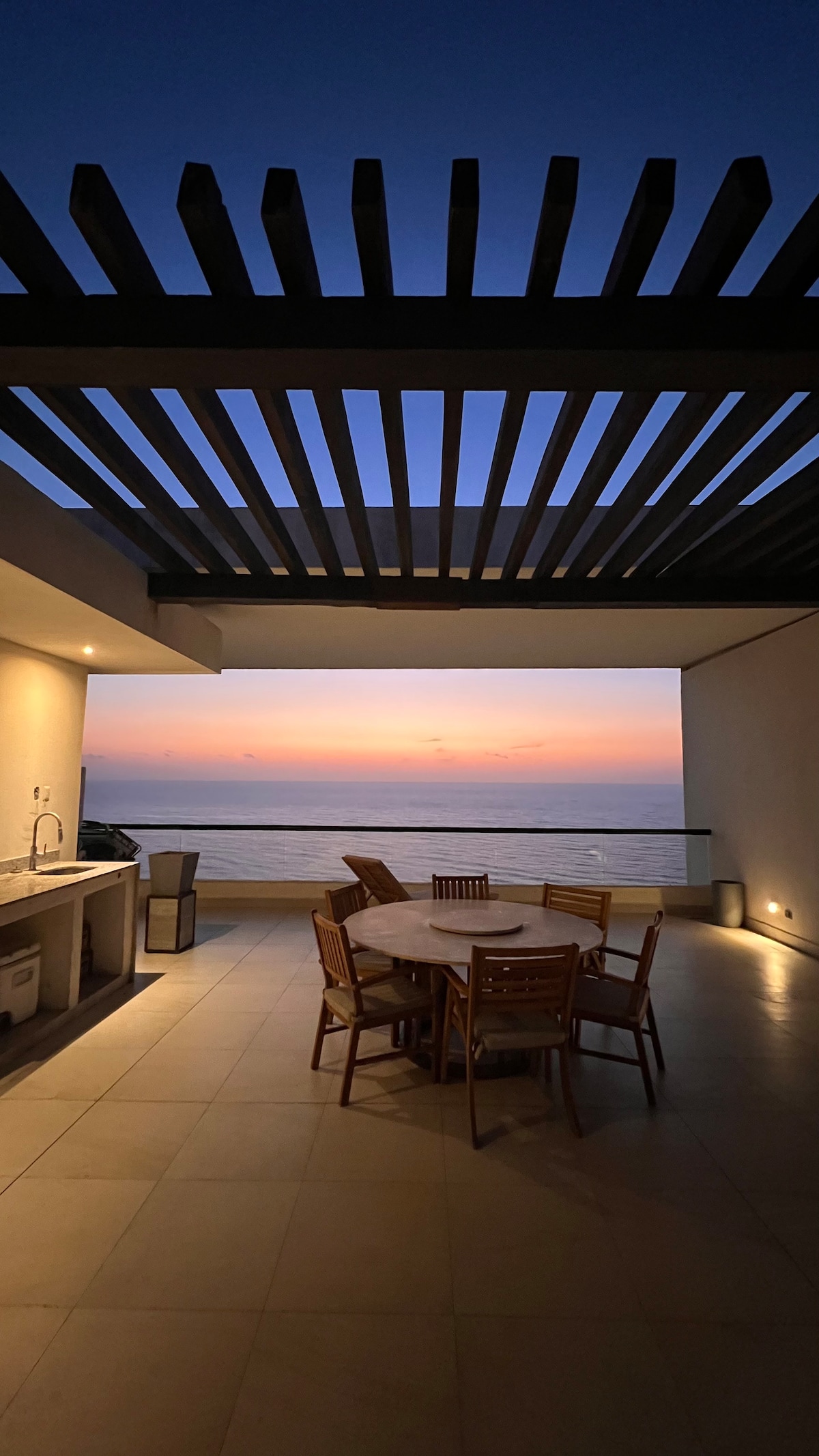 An outdoor dining area is featured, with a round table surrounded by six wooden chairs. A built-in grill is positioned to the side, and the expansive ocean view is framed by slatted overhead beams. Warm sunset colors illuminate the scene.