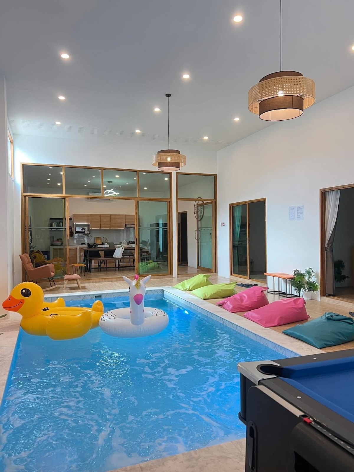 An indoor pool area features colorful floating toys, including a large yellow duck and a white unicorn. Bright bean bags in shades of pink and green are arranged nearby. Natural light illuminates the space through expansive glass doors leading to a visible common area.