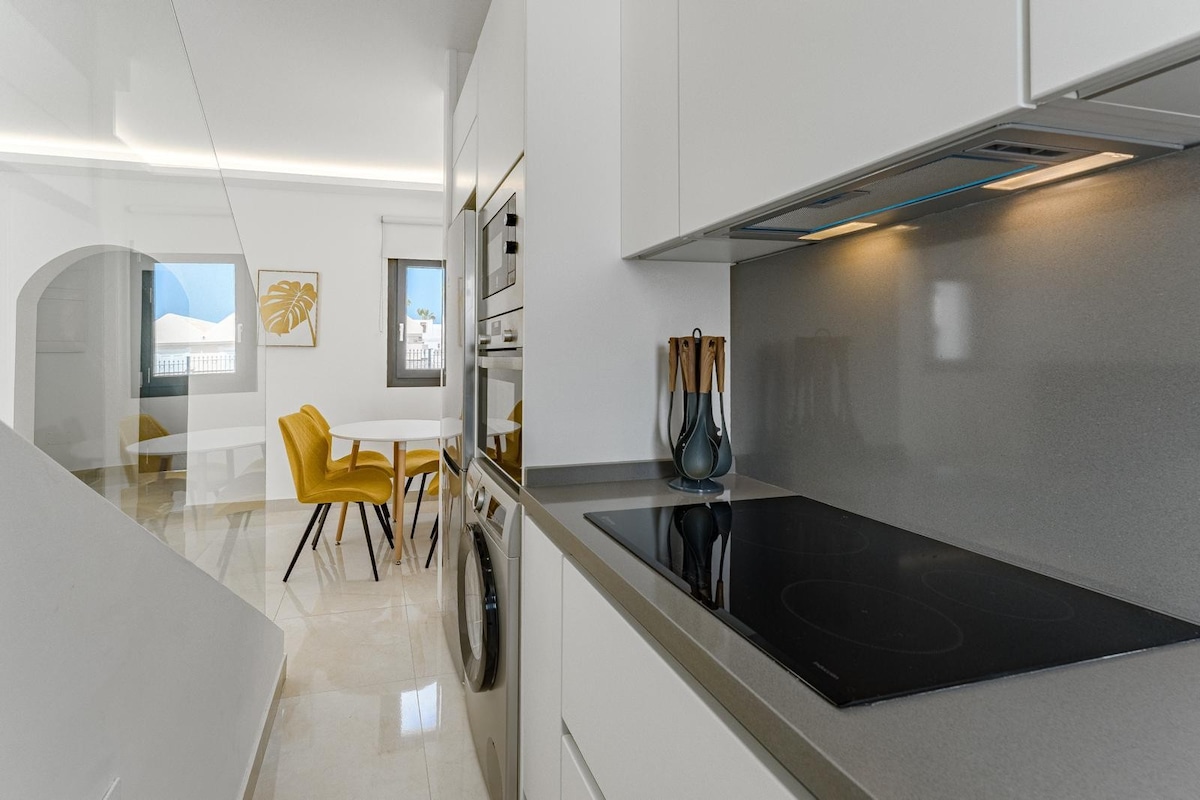 A modern kitchen area features sleek gray countertops and built-in appliances, including a cooktop and washing machine. A dining table with two yellow chairs is positioned near a window, providing ample natural light to the space.
