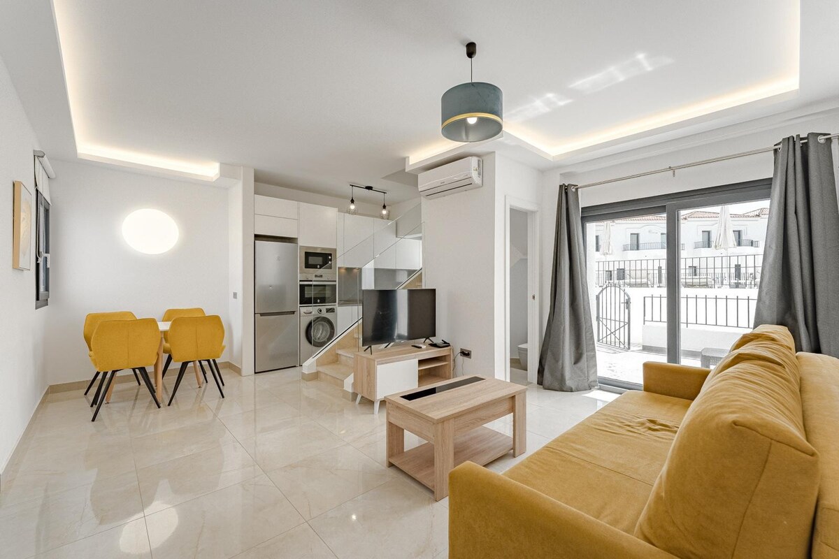 A modern living area features a yellow sofa facing a flat-screen television. Light-colored tiles reflect natural light. A dining space with two chairs is positioned nearby, while large glass doors provide access to an outdoor area. The space is complemented by neutral-toned decor.