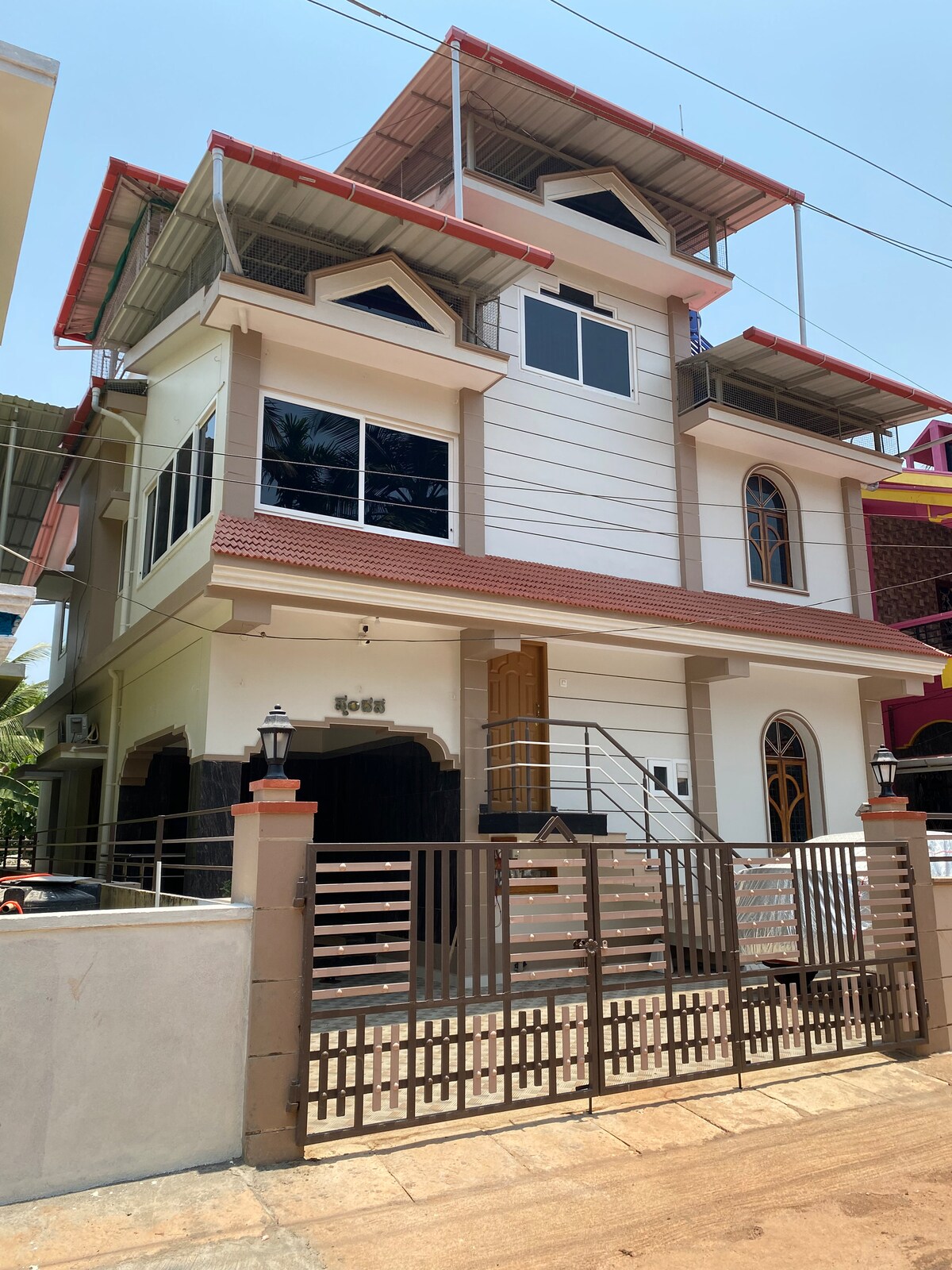 The exterior of a two-story house is depicted, featuring a mix of modern and traditional architectural elements. Large windows allow for ample natural light, while a covered entrance adds functionality. The front gate is securely designed, surrounded by a well-maintained pathway.