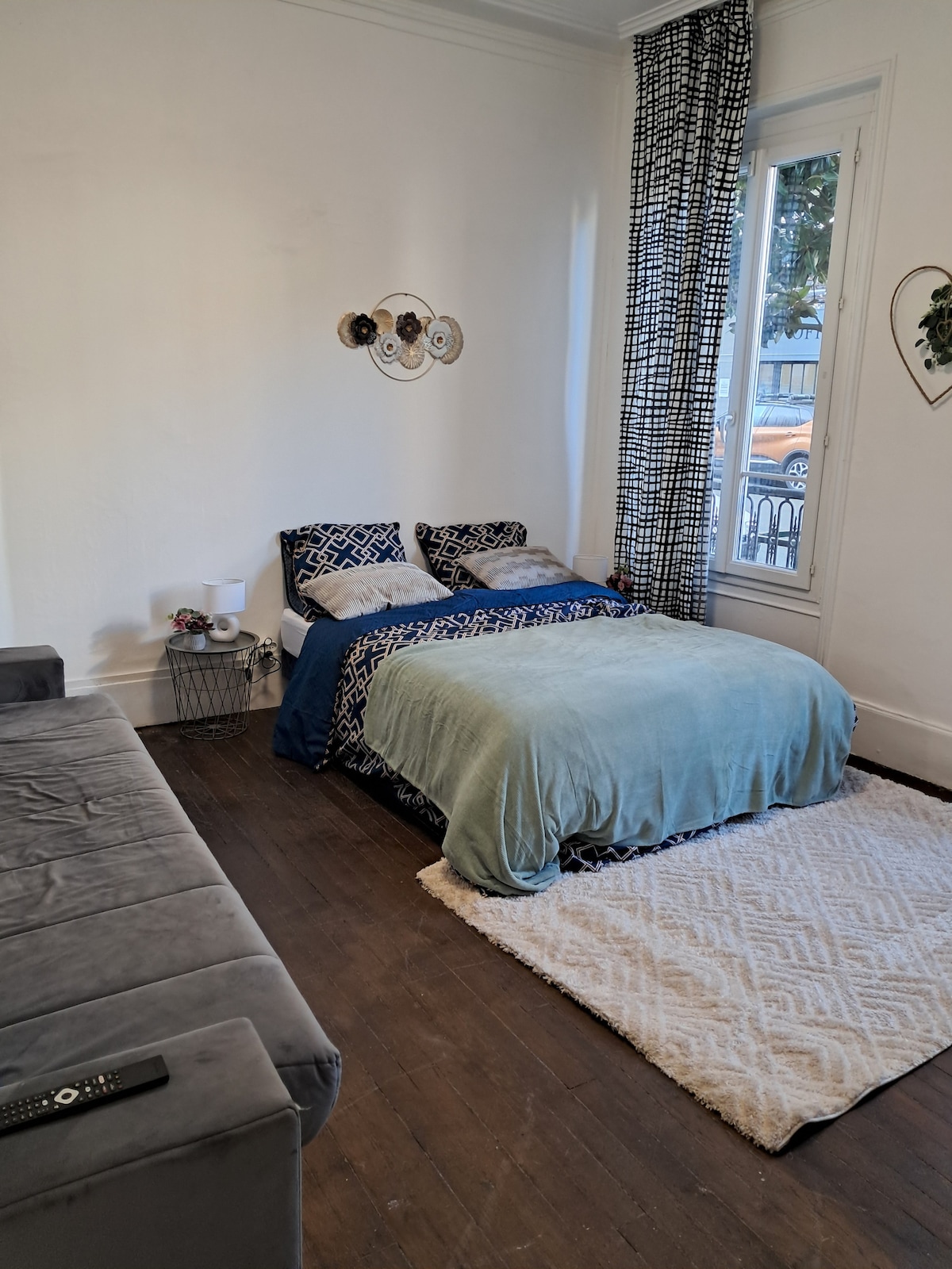 A cozy sleeping area is presented, featuring a spacious bed adorned with a blue and white patterned coverlet. A soft green throw lies across the bed, complemented by coordinating pillows. Natural light filters through a nearby window covered by a sheer curtain, illuminating the wooden floor.