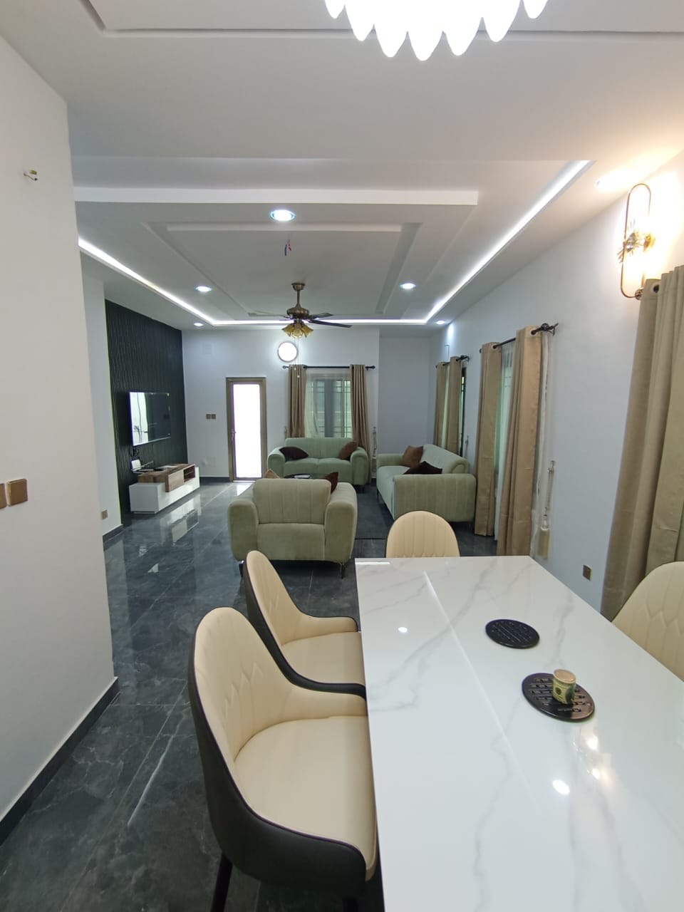 An open living and dining area is presented, featuring a marble-top dining table accompanied by beige chairs. Soft green seating arrangements are positioned nearby, complemented by large windows with curtains allowing natural light to enhance the elegant décor. A flat-screen TV is mounted on one wall.