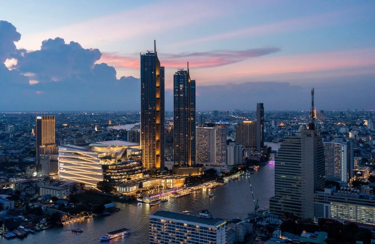 An aerial view captures the vibrant skyline of Bangkok during twilight, showcasing several illuminated skyscrapers along the Chao Phraya River. The scene reflects a blend of modern architecture and the dynamic city life, enhanced by soft hues from the setting sun.