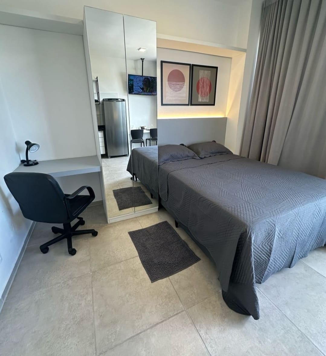 A modern bedroom features a spacious bed dressed in grey linens, complemented by neutral-toned curtains and a large mirror. A workspace is visible with a desk and a black office chair. Soft, patterned rugs are positioned on the tiled floor.