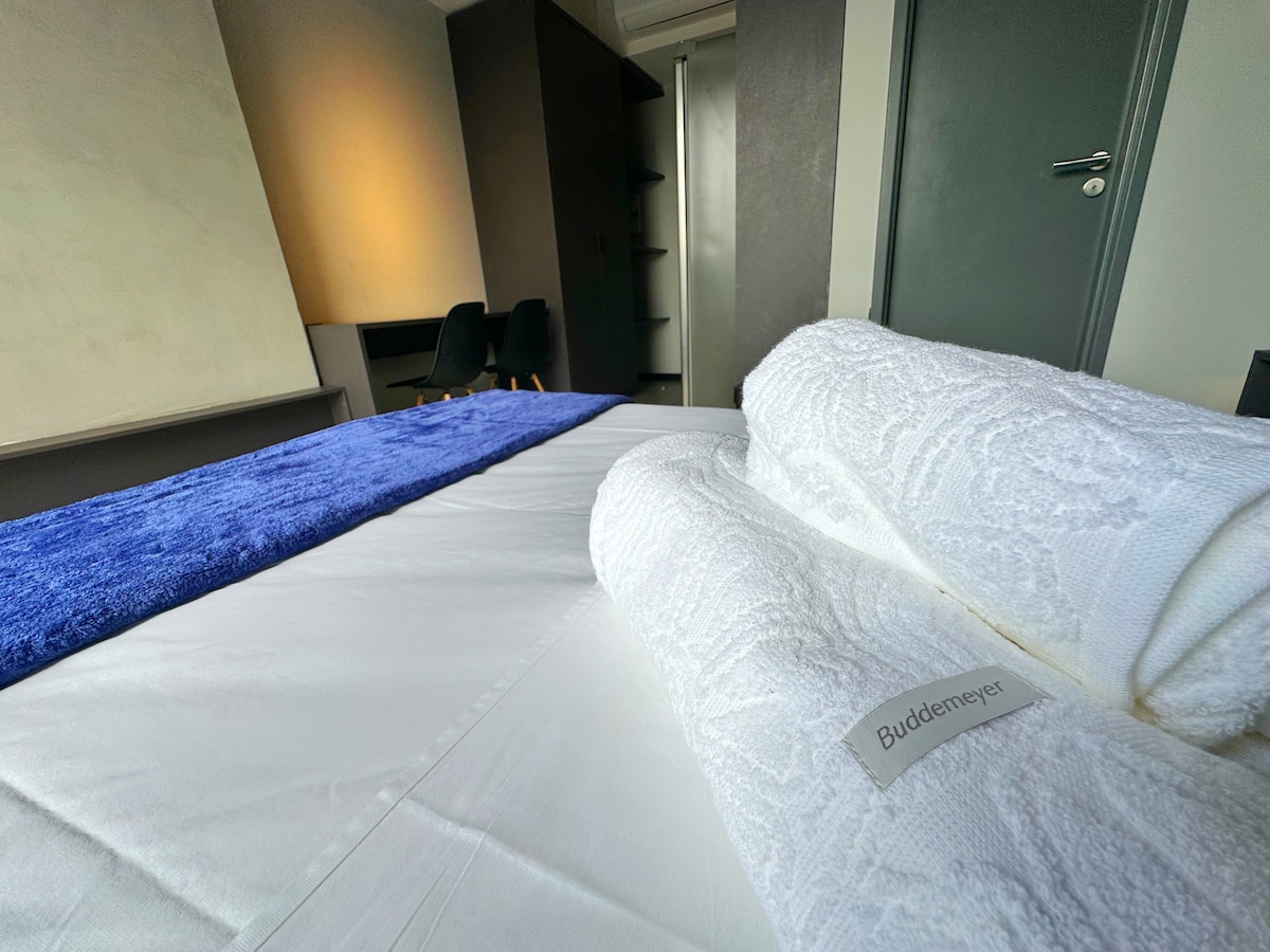 A neatly arranged bed features a crisp white sheet complemented by a blue throw blanket. Soft, rolled towels are placed at the foot of the bed. The background reveals minimalistic furniture and a wardrobe, with natural light softly illuminating the space.