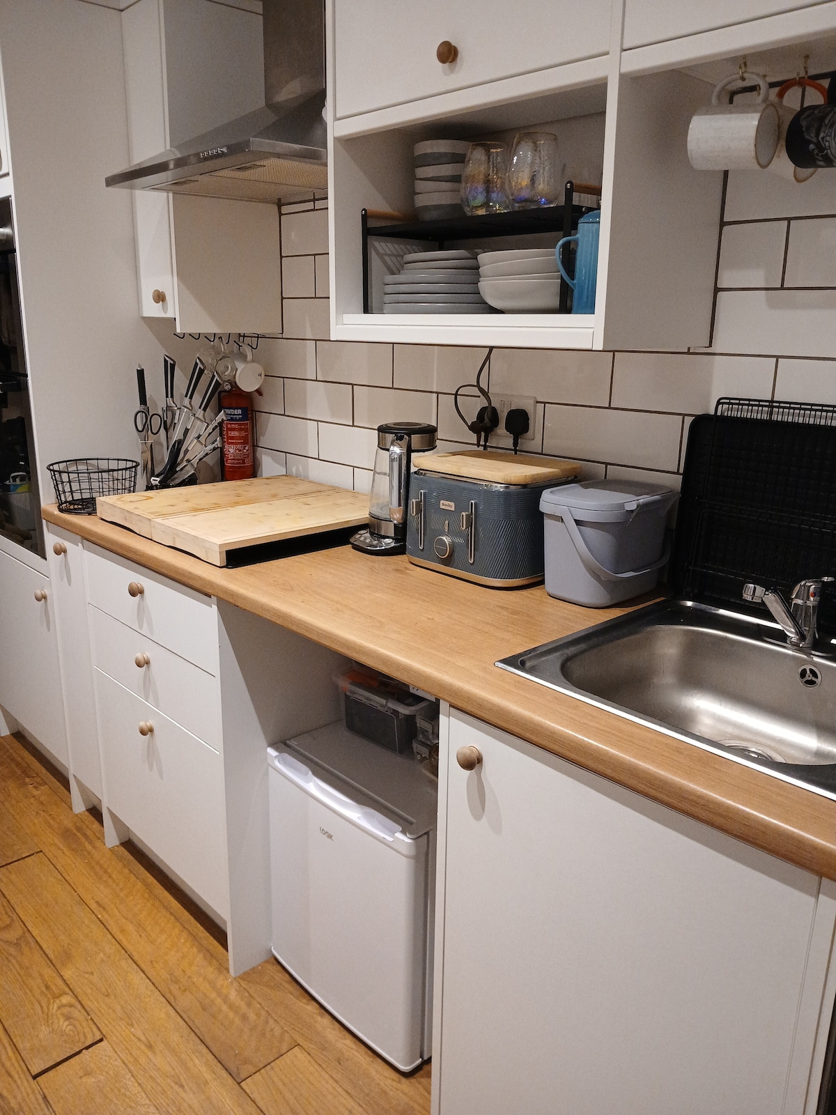 The compact kitchen features sleek white cabinetry and a warm wooden countertop. Essential appliances, such as a double oven, grill, and small under-counter freezer, are included. Utensils and dishware are neatly organized, enhancing the functional design of the space.