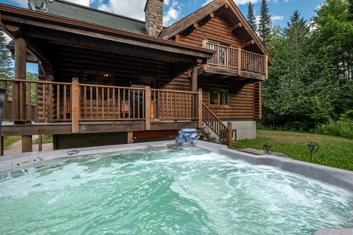 A spacious outdoor spa is positioned in the foreground, with water bubbling invitingly. The log cabin, characterized by its natural wood exterior, stands in the background, bordered by lush greenery. A wooden deck and balcony are visible, enhancing the rustic charm.
