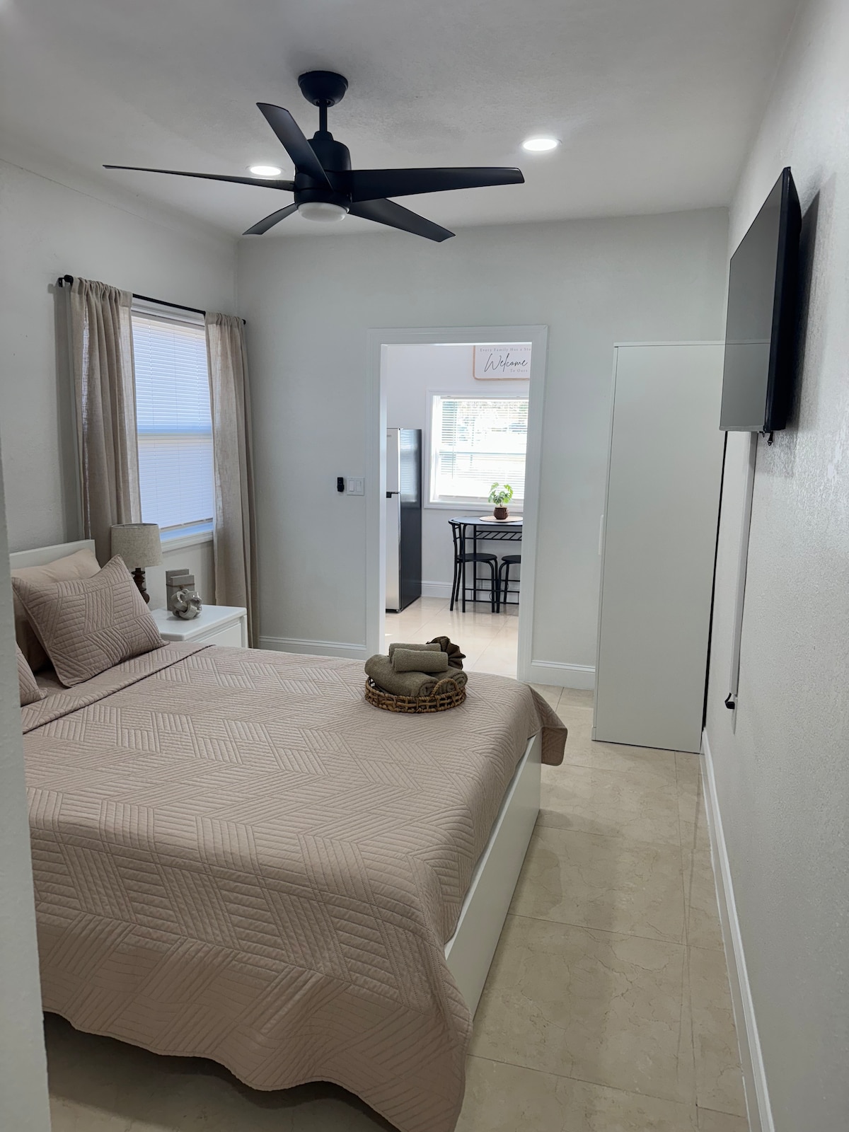 A serene bedroom features a spacious bed with a light beige quilt. A ceiling fan is positioned overhead, and natural light enters through a nearby window. A small side table holds decorative items. An open doorway leads to an adjoining area with a dining table.
