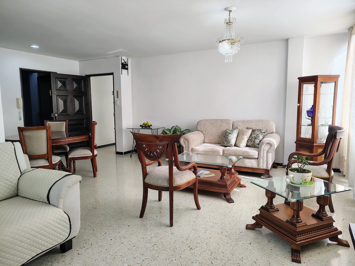 A spacious living area features a mix of seating options, including a beige sofa with patterned cushions, wooden armchairs, and a glass coffee table atop a light-colored floor. Natural light enters through large windows, enhancing the inviting atmosphere.