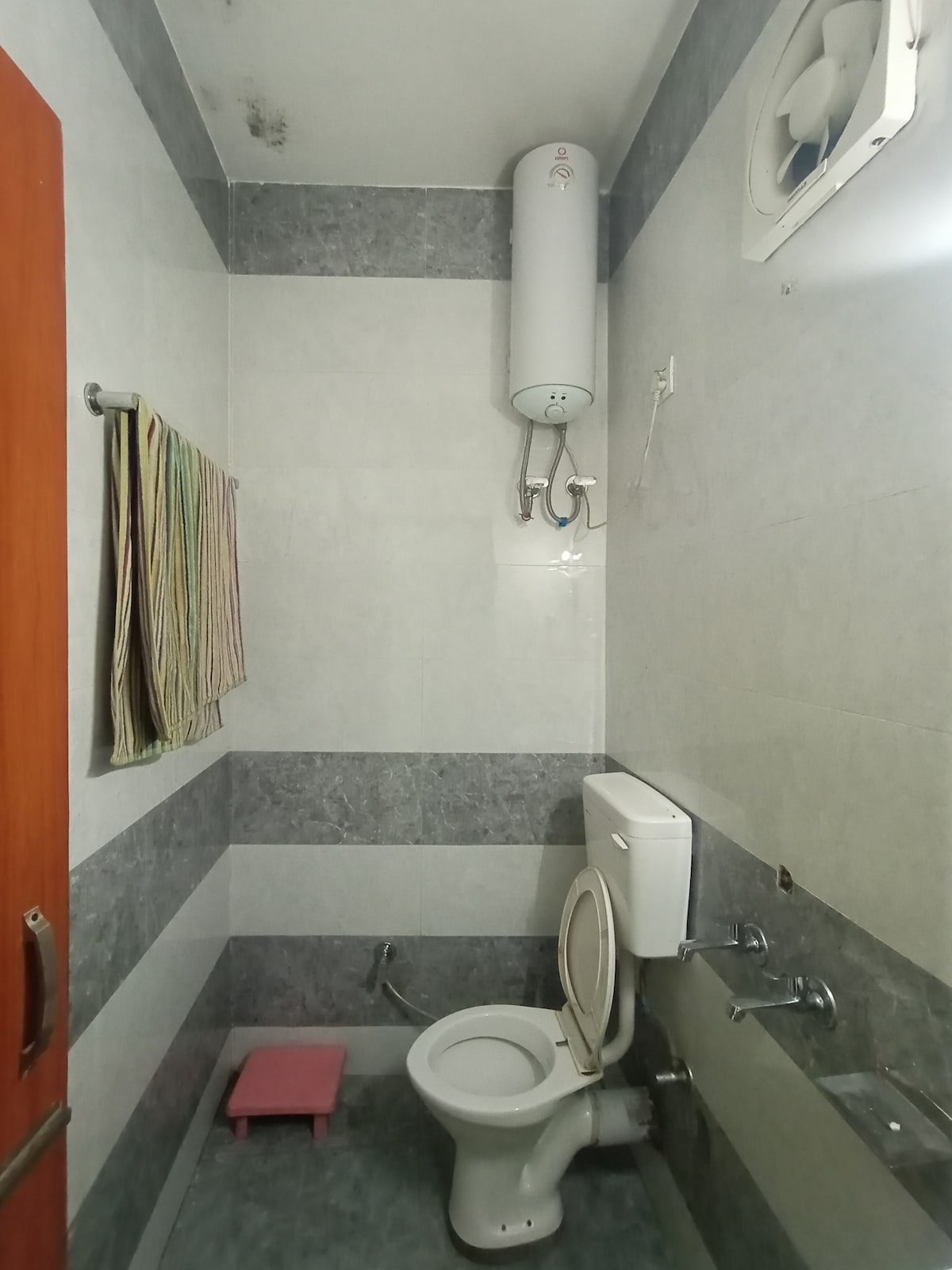 A compact bathroom is equipped with a toilet and a wash basin, set against light and dark grey tiled walls. A small pink stool is placed on the floor. A water heater is mounted above, alongside a vent for airflow. Several towels are neatly hung on a rack.