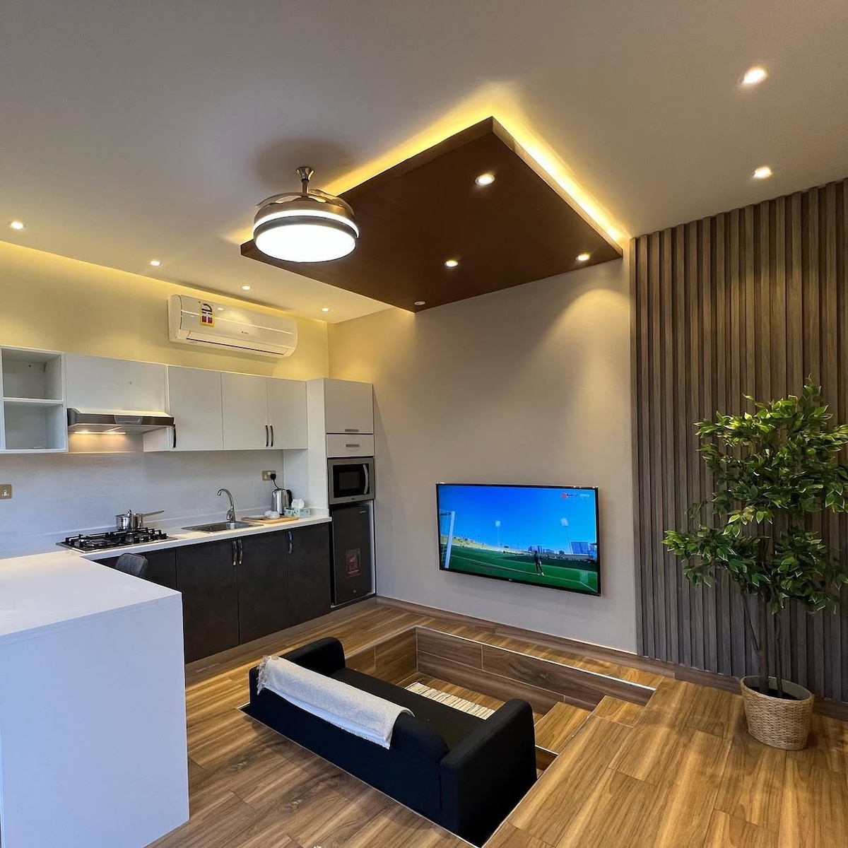 A modern living space is presented with an open kitchen featuring sleek cabinetry and essential appliances. A minimalist black sofa is positioned in front of a mounted television, while a potted plant adds a touch of greenery. Soft lighting enhances the overall ambiance.