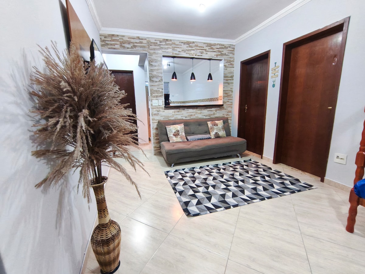 A spacious living area features a large, patterned rug positioned beneath a cozy sofa, with decorative cushions adding a touch of comfort. The walls are adorned with light-colored finishes, and three pendant lights hang above a mirror, reflecting the warm ambiance of the space.