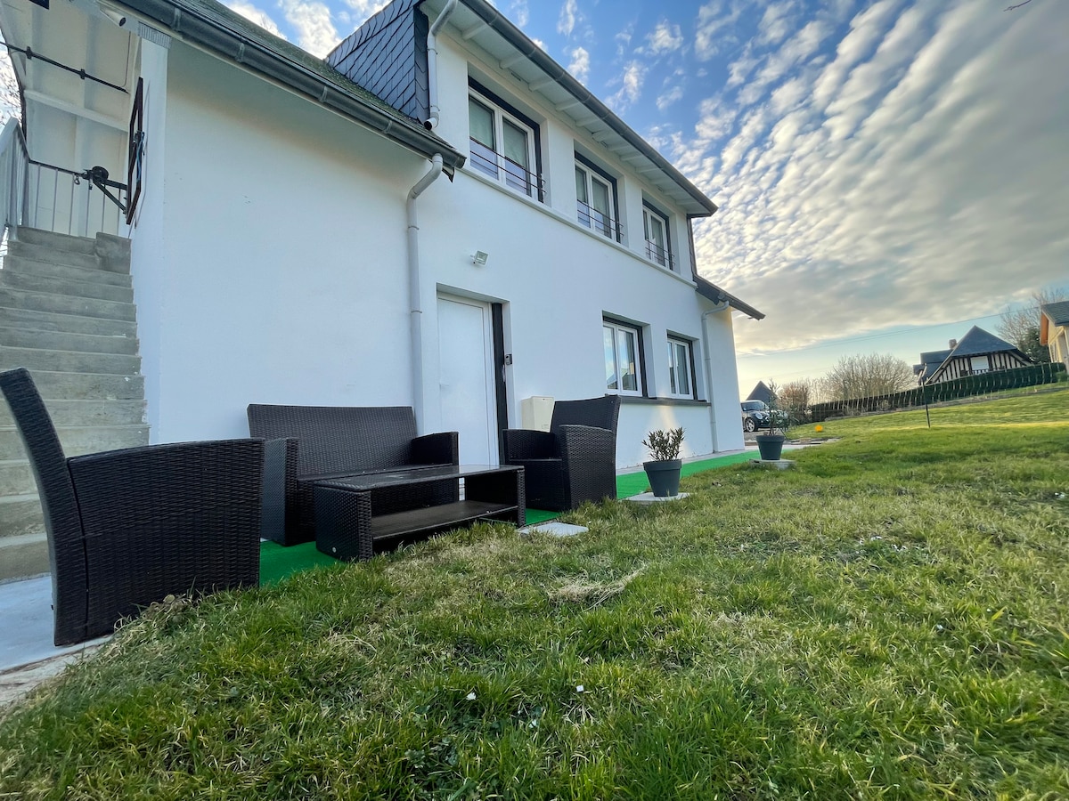 An outdoor area features a grass lawn with modern outdoor furniture arranged for relaxation. A small table is complemented by four wicker chairs. The white exterior of the building is visible, along with nearby steps leading to the entrance, under a sky filled with scattered clouds.