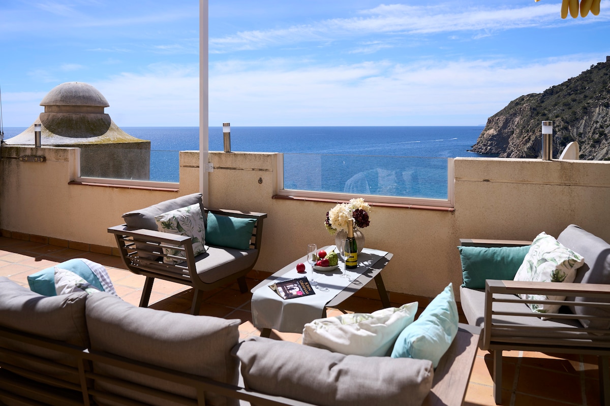 A spacious terrace features modern outdoor furniture, including a sectional sofa and a coffee table. Soft cushions in light tones are placed on the seating. The panoramic sea views stretch across the horizon, complemented by the natural landscape in the background.