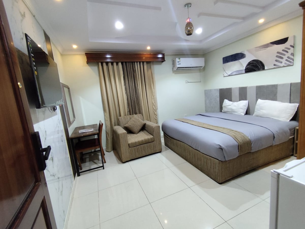 A spacious room features a large bed with neutral-toned bedding, complemented by two pillows. A cozy armchair is situated near a compact desk, while an LCD television is mounted on the wall. Natural light illuminates the space, creating a welcoming ambiance.