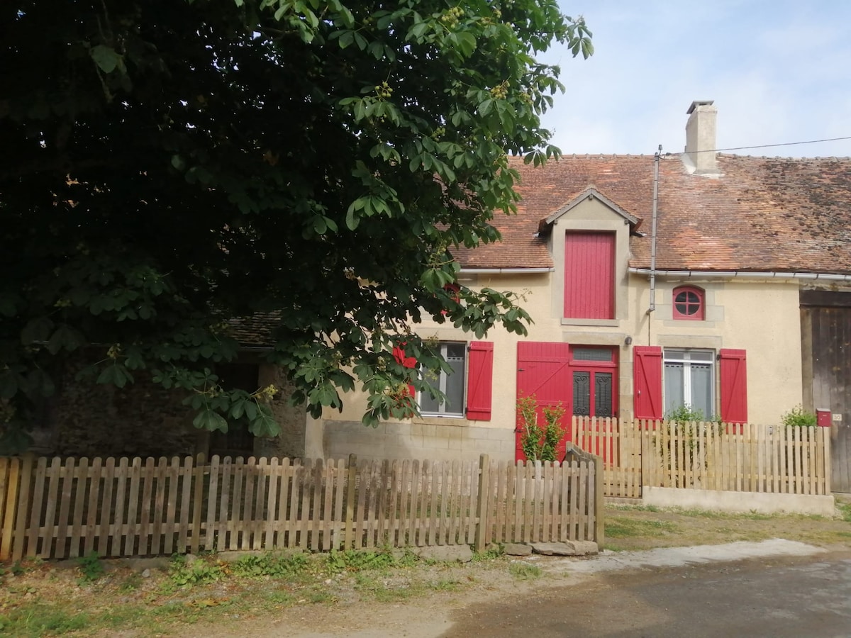The charming exterior of a rural house is visible, featuring rustic stone walls and red shuttered windows. A white picket fence encloses the front yard, providing a welcoming boundary. Lush greenery surrounds the home, enhancing its quaint appearance against a clear sky.