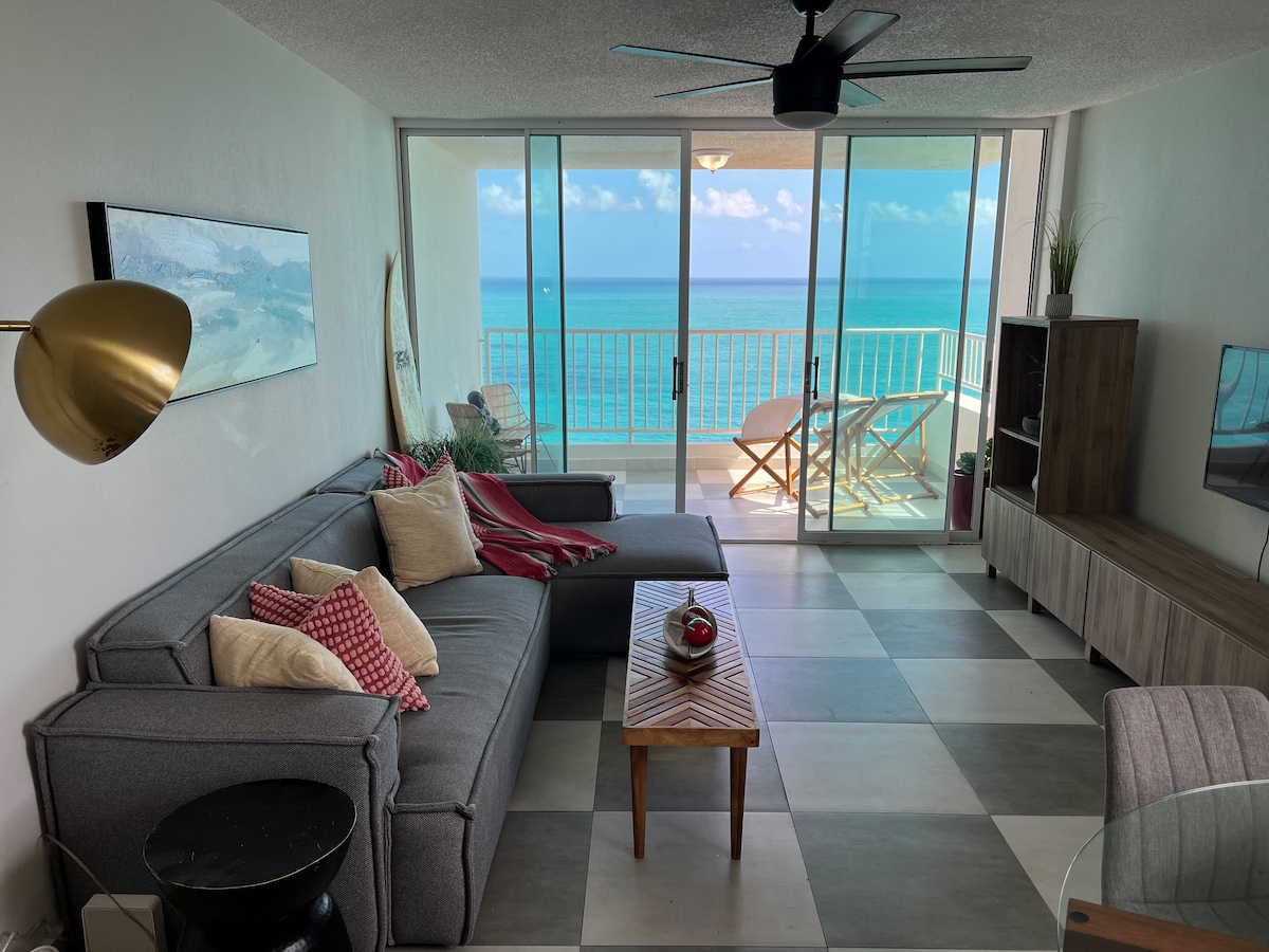 A modern living area is bathed in natural light from the large glass doors, which offer panoramic ocean views. A spacious gray sofa is paired with a wooden coffee table, while a ceiling fan provides a comfortable atmosphere. The room features decor elements and subtle accents that invite relaxation.