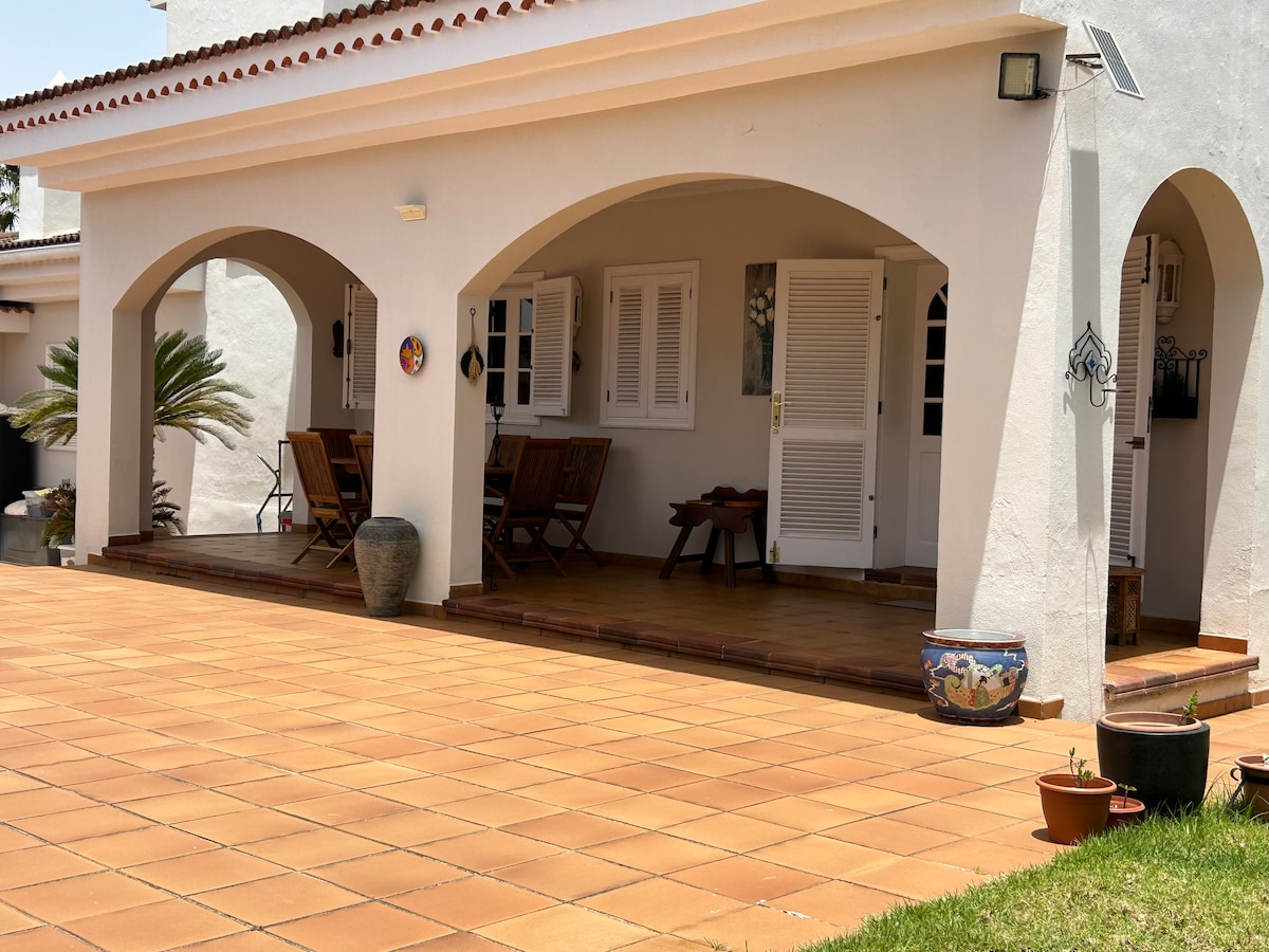 A shaded terrace features wooden furniture arranged for outdoor seating, complemented by potted plants and decorative elements. The tiled floor extends to the entrance, showcasing a welcoming space that blends seamlessly with the surrounding greenery.