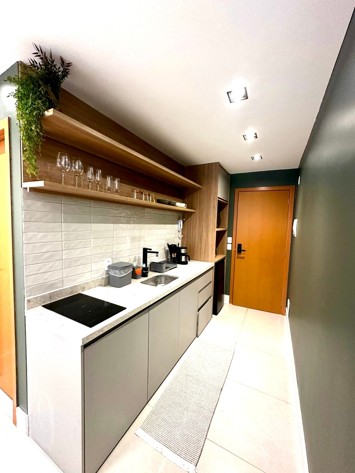 A compact kitchen area features sleek cabinets and light-colored countertops. Above, open shelves display glassware, while various appliances are neatly arranged. Natural light from a nearby door illuminates the space, complemented by a textured rug on the floor.