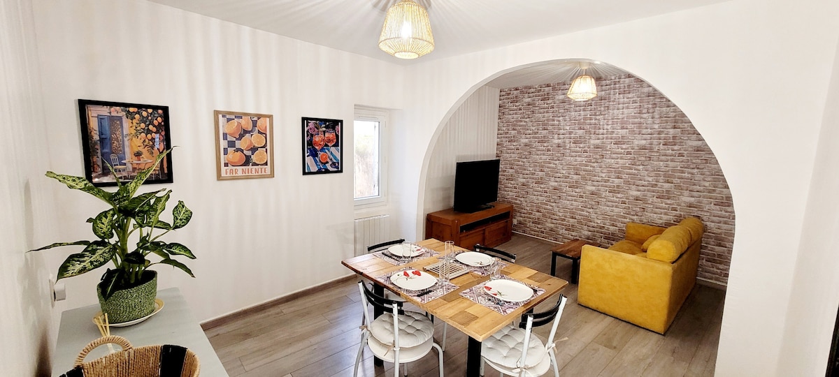 A dining area is positioned within an open-plan space, featuring a wooden table set for four. Light streams through a window, illuminating the room's soft tones. On one side, a comfortable yellow sofa sits against a textured wall, creating a cozy atmosphere. Decorative artwork adorns the walls.