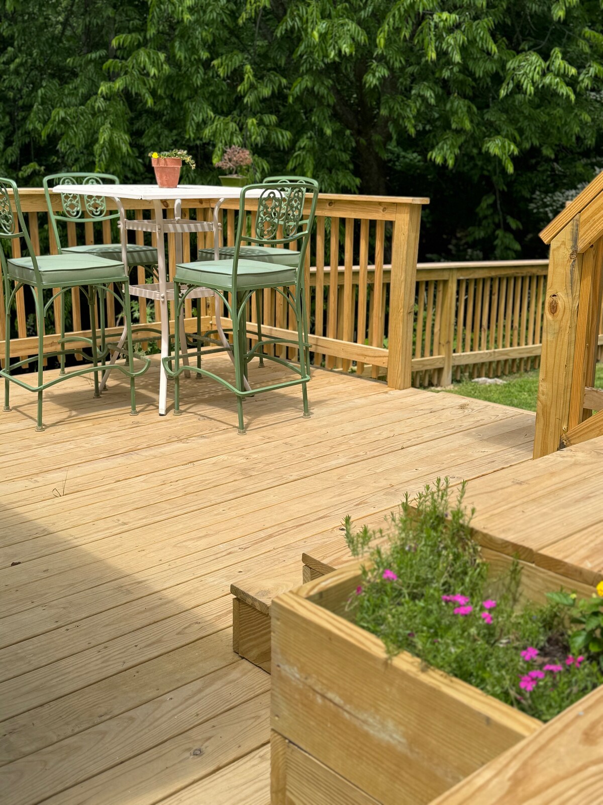 A wooden deck features a small round table surrounded by two green bar stools, providing an ideal spot for outdoor relaxation. A planter box filled with vibrant flowers adds a touch of color, while trees create a natural backdrop, enhancing the tranquil setting.