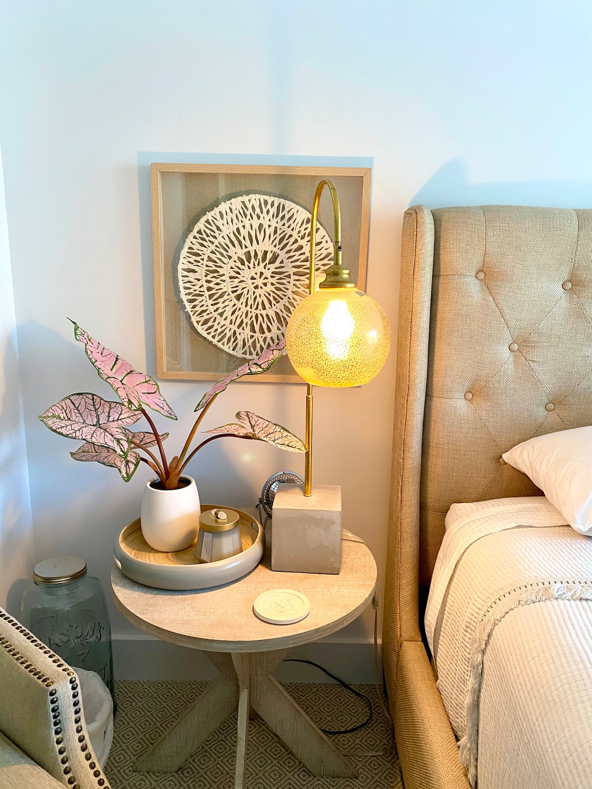 A bedside table features a textured lamp with a warm glow, alongside a decorative plant and a small tray. The background includes an art piece with a circular design. The neutral color scheme creates a calming atmosphere in the cozy sleeping area.