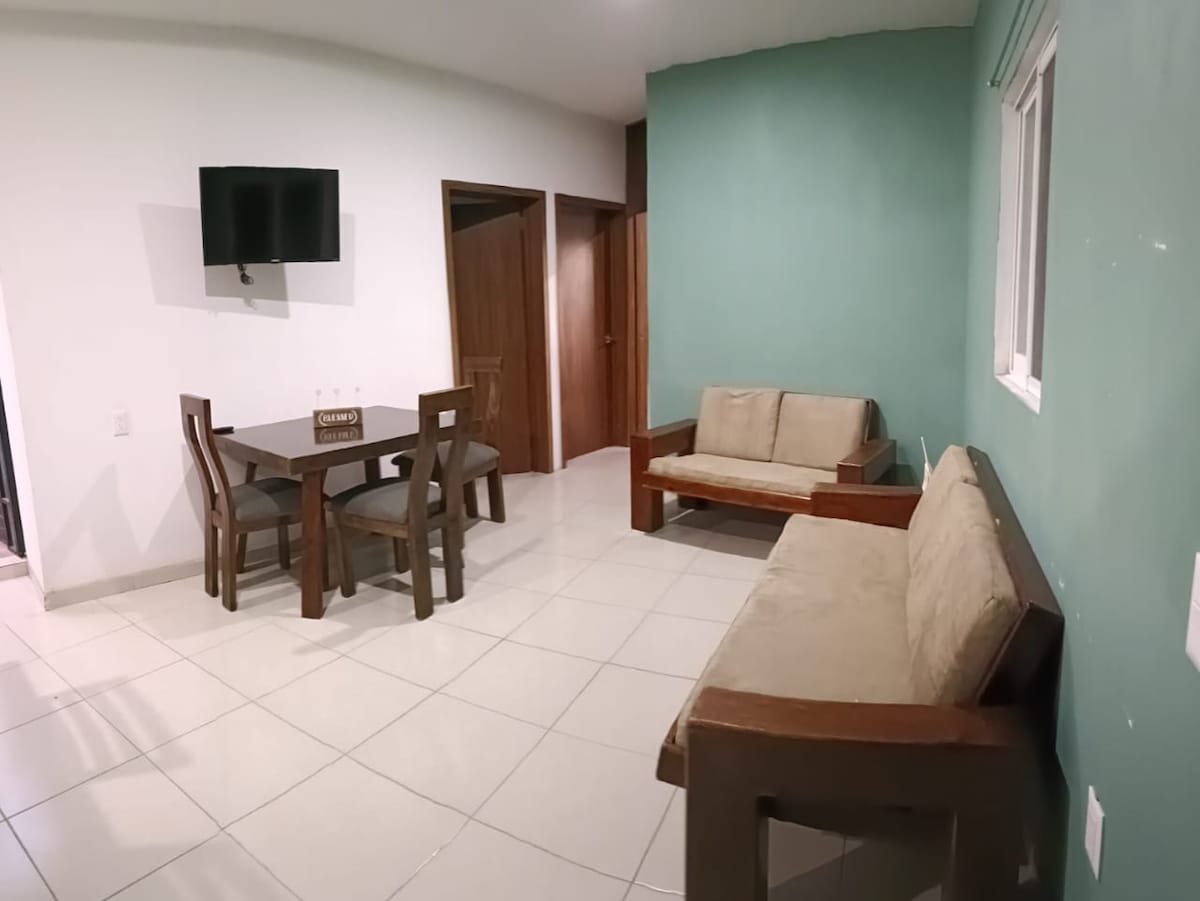A spacious living area is equipped with a wooden table surrounded by four chairs. A comfortable sofa and a loveseat offer seating options. A wall-mounted television is visible, and light enters through a nearby window, which is partially covered.