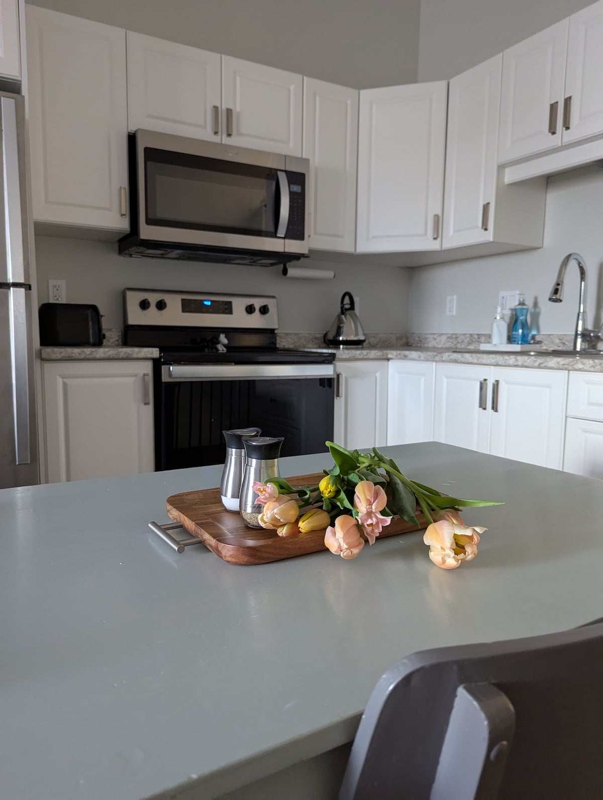 A modern kitchen is shown, featuring stainless steel appliances and light cabinetry. A countertop is adorned with a wooden tray holding a small coffee pot and a bouquet of fresh flowers, adding a subtle touch of color to the space.