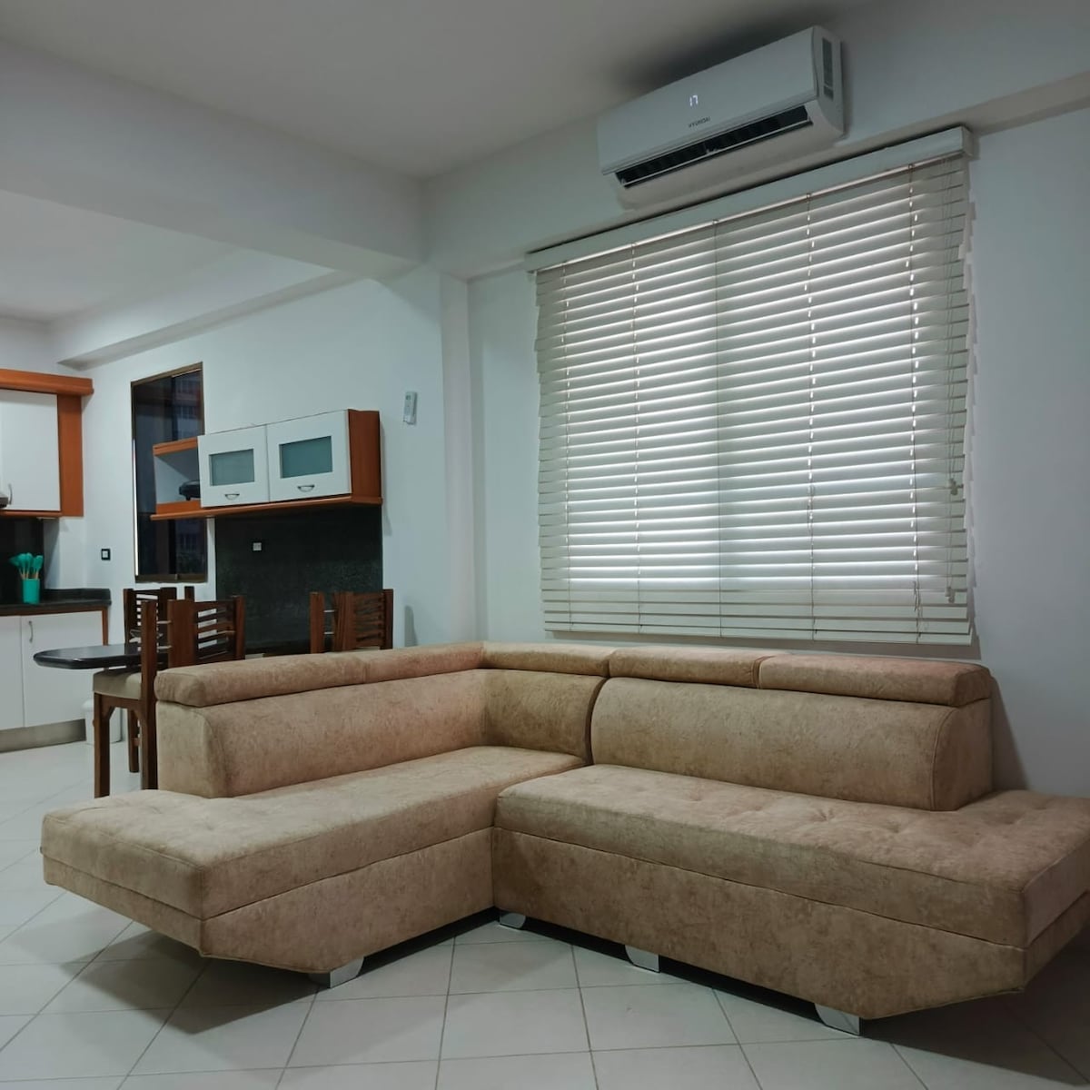 A cozy living area features a spacious sectional sofa in a light beige fabric, complemented by a sleek dining table with four chairs in the background. Large windows with horizontal blinds allow natural light to brighten the space.