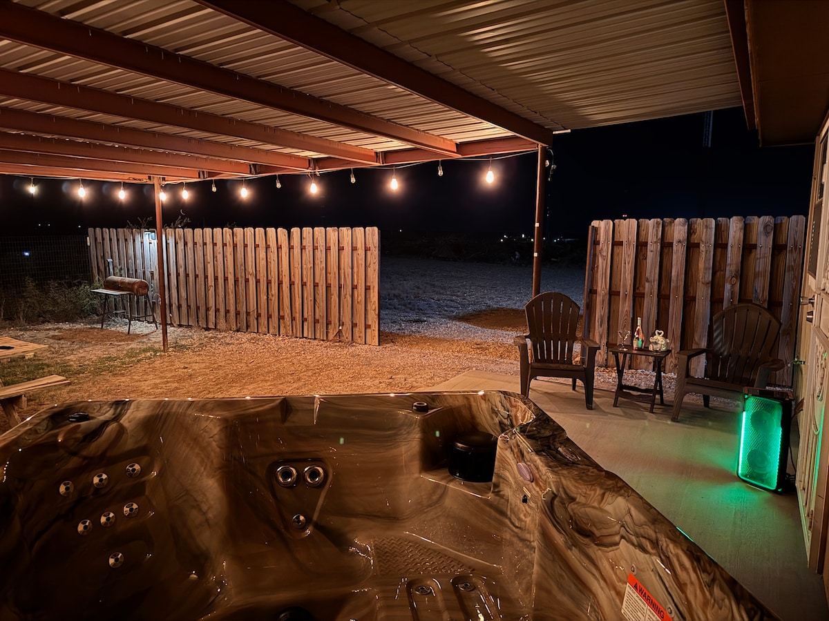 The heated jacuzzi is positioned under a covered area, surrounded by a wooden fence. Soft ambient lighting strings across the ceiling, creating a serene atmosphere. Nearby, two chairs are placed on a concrete surface, with a small table featuring decorative items illuminated by a soft glow.