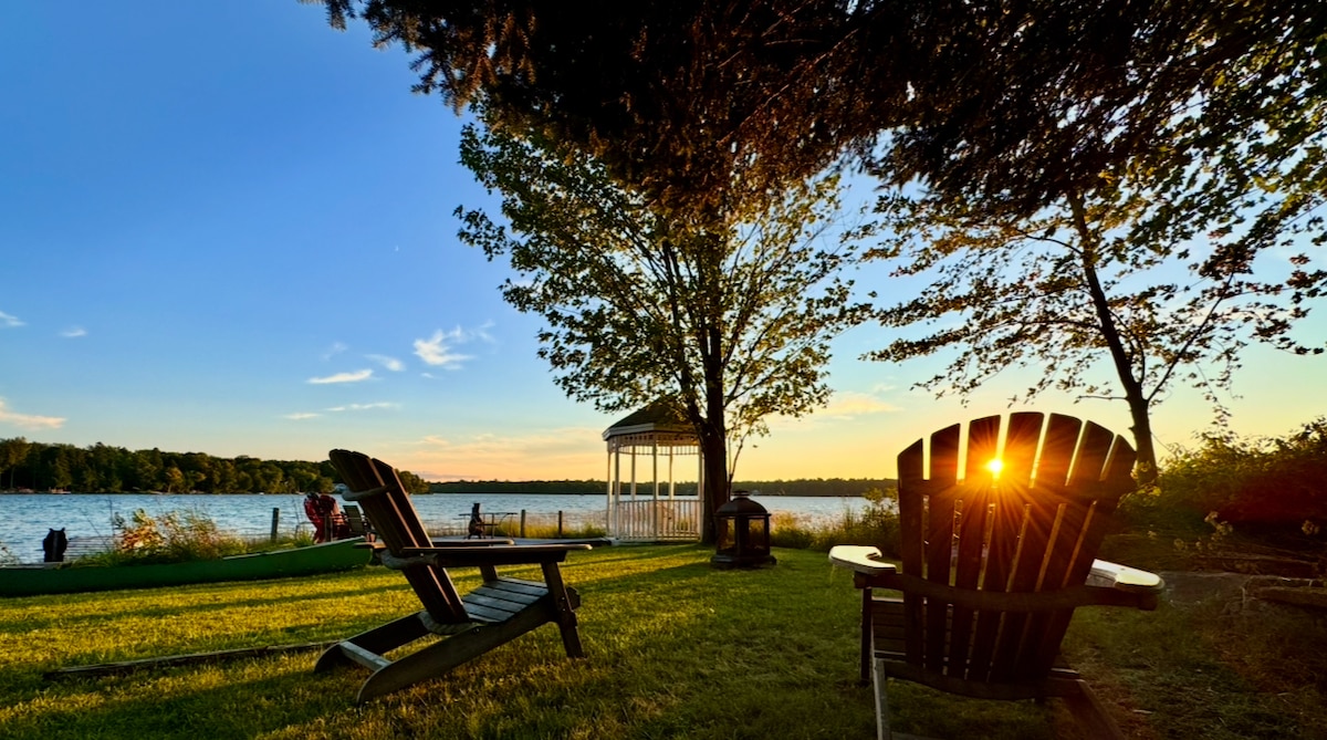 The image features a serene waterfront view with two wooden Muskoka chairs positioned on green grass. A gazebo can be seen in the background beside the water, with the sun setting on the horizon, casting a warm glow across the scene.