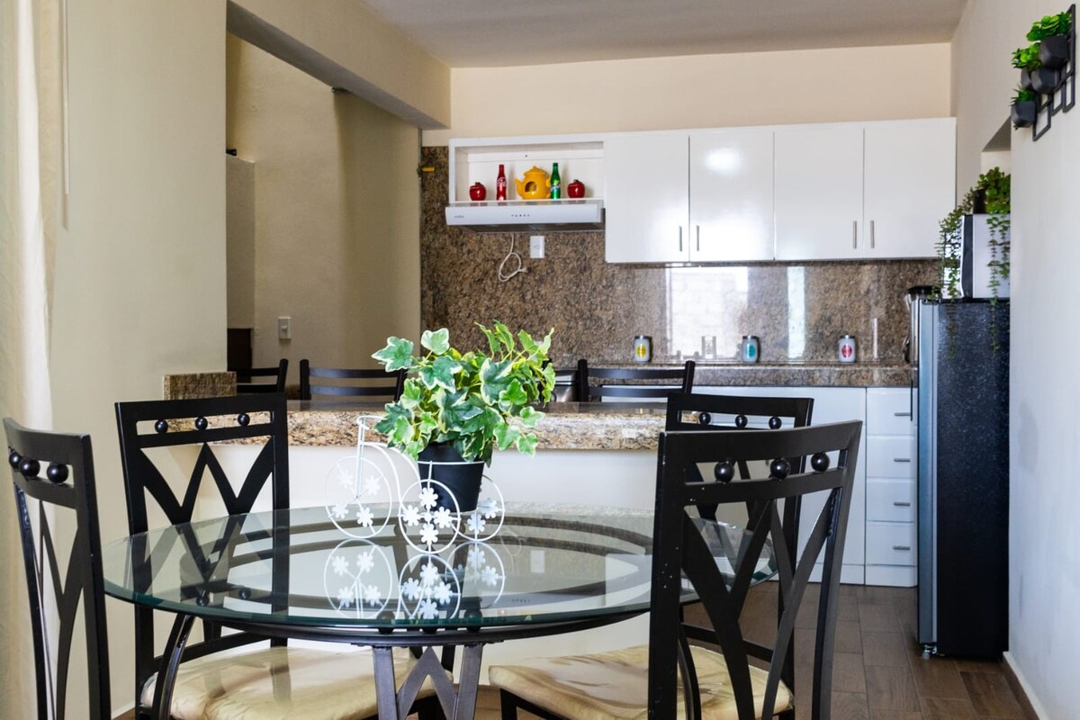 A modern kitchen area features sleek white cabinetry and a granite countertop. A round glass dining table surrounded by four black chairs is positioned near the kitchen, with a potted plant adding a touch of greenery.