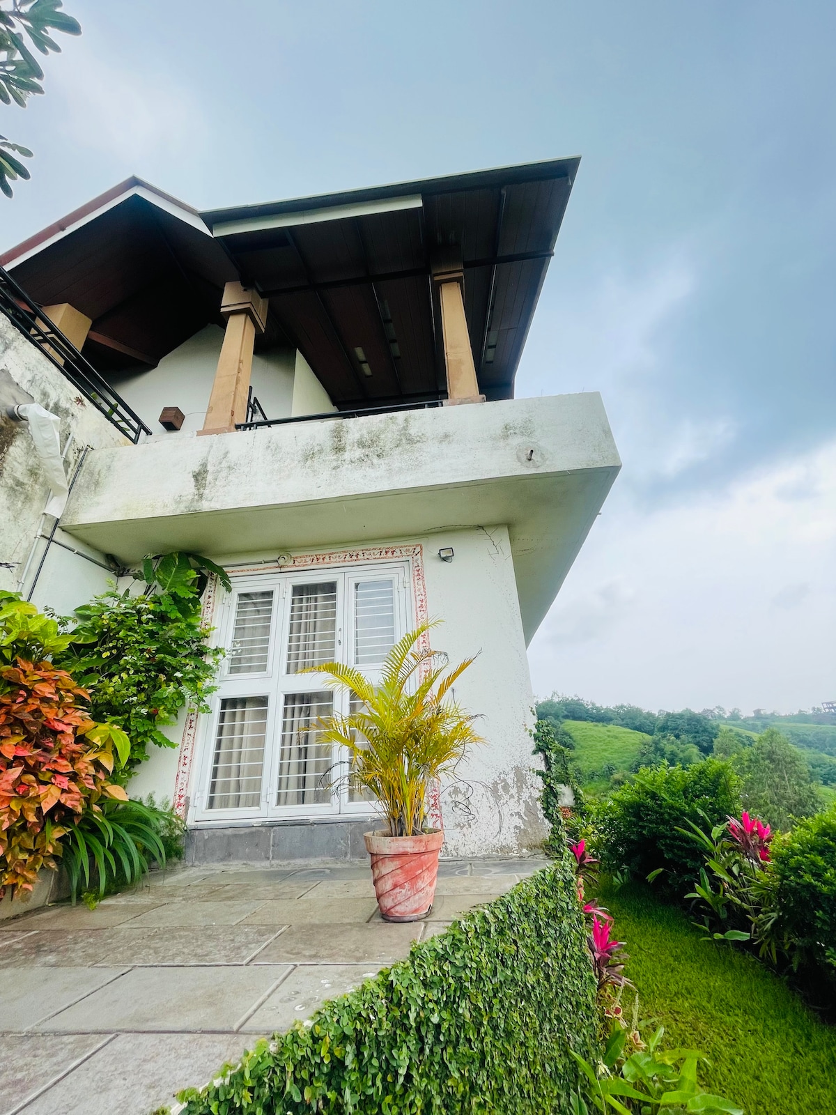 The exterior of the villa is showcased from a low angle, featuring a modern roof overhanging a small balcony. Lush greenery and vibrant plants frame the entrance, while a terracotta pot with a fern adds a touch of nature. Scenic hills are visible in the background.