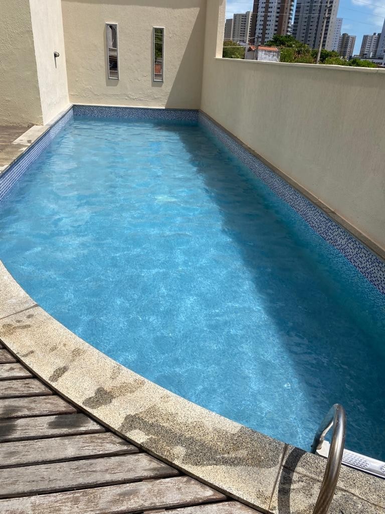 A tranquil swimming pool is shown, featuring clear blue water and a textured stone edge. Sunlight reflects off the surface, creating a serene atmosphere. The pool is bordered by smooth walls and overlooks a cityscape in the background.