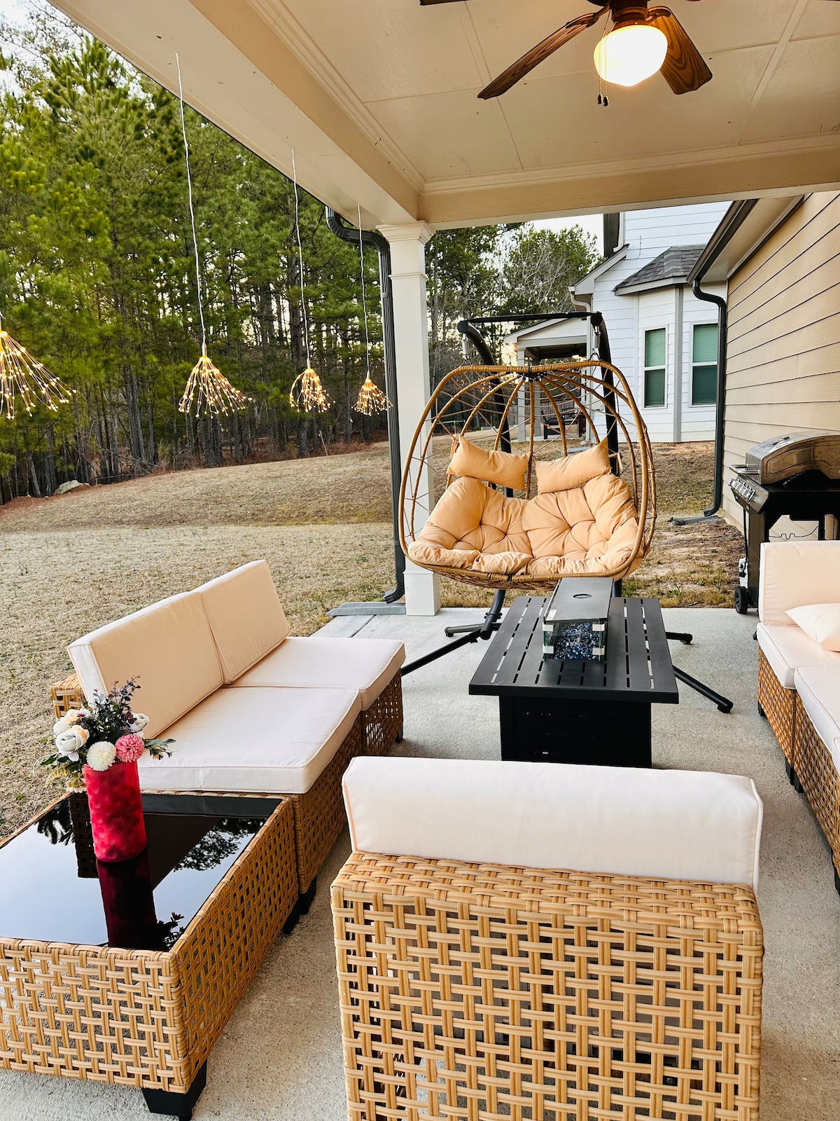 A cozy outdoor seating area is presented, featuring beige cushioned sofas arranged around a black fire table. A hanging chair with cushioned seating adds an inviting touch. The setting is complemented by decorative lighting suspended above, with a natural area of grass and trees visible in the background.