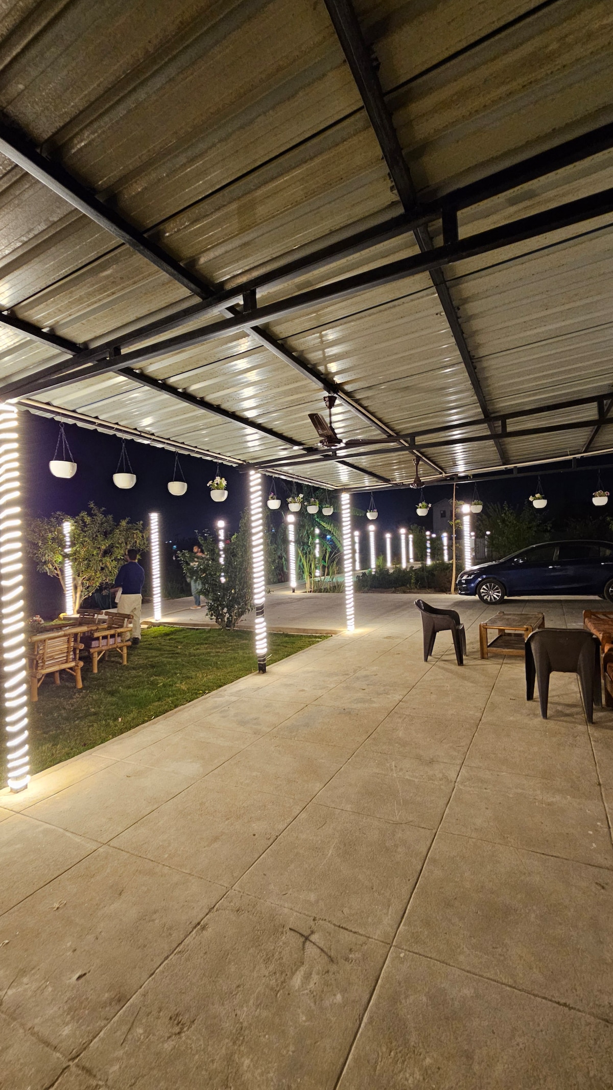 An outdoor seating area is illuminated by soft lighting, creating a serene ambiance. The space features wooden seating arrangements under a covered patio, with greenery visible in the background. Unpaved flooring provides easy access to the comfortable chairs and tables placed throughout.