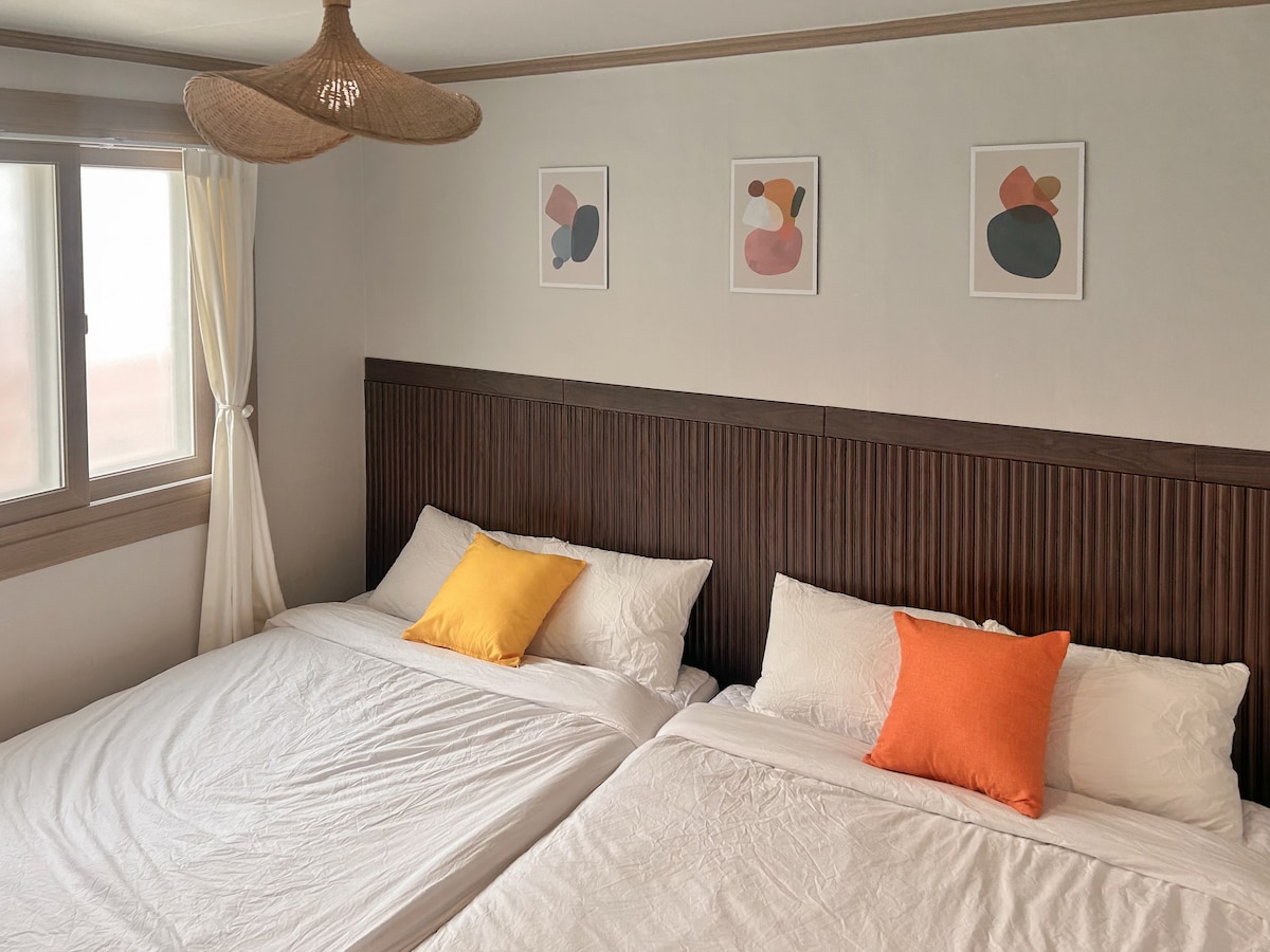A serene bedroom is displayed, featuring two spacious beds dressed in crisp white linens. Bright cushions in shades of yellow and orange add a pop of color. Above the beds, minimalist artwork in soft tones is arranged on the wall, enhancing the calm ambiance.