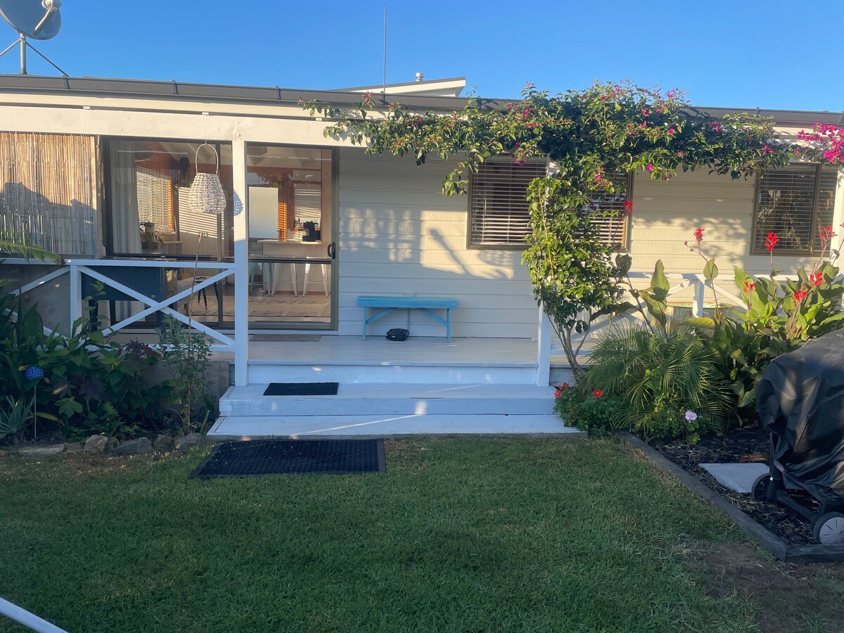 A welcoming entrance is displayed, featuring a small blue bench on a white-painted porch. Lush greenery and colorful flowers frame the walkway, which leads to a glass door that opens to the interior. A grill is positioned nearby, indicating a space for outdoor cooking.