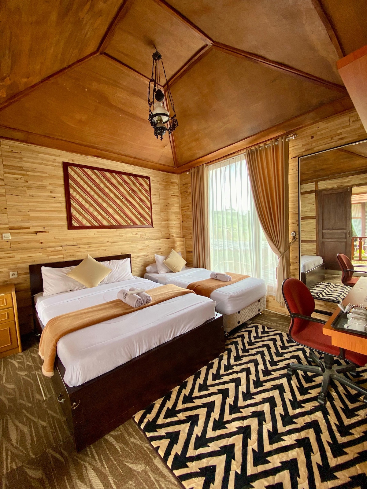 A cozy guest room is presented with two spacious beds dressed in crisp white linens. Natural wood paneling and a high, sloped ceiling enhance the interior. A patterned rug covers the floor, while large windows with sheer curtains allow soft light to fill the space. A desk and chair are situated by the mirror.