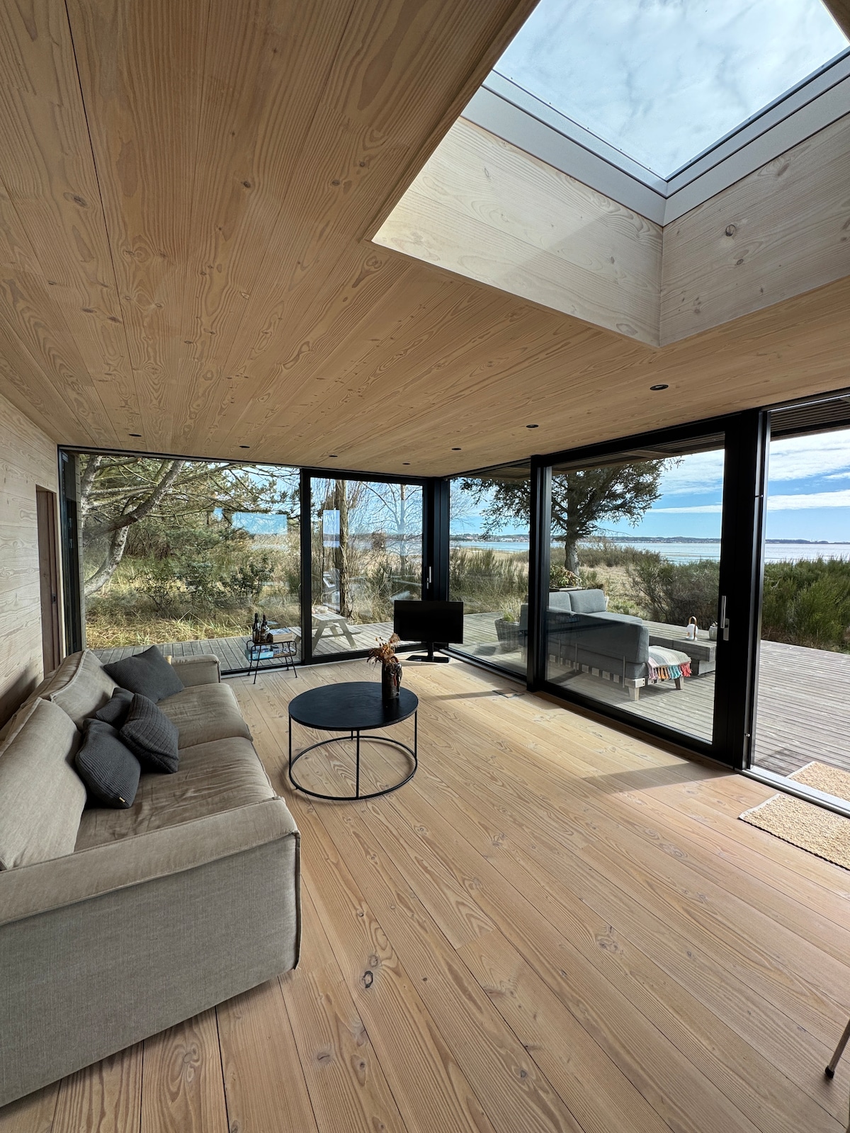 An airy interior room features large glass panels that provide expansive views of the natural surroundings. A cozy sofa is paired with a round coffee table, and a flat-screen TV is visible in the corner. A skylight allows natural light to fill the space.