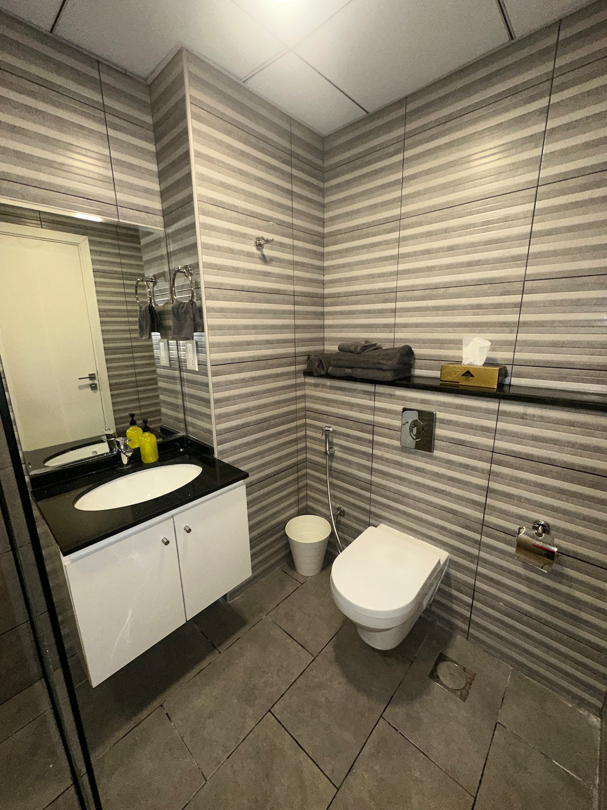 The modern bathroom features a sleek design with gray and white striped walls. A large mirror is positioned above a black countertop with a circular sink. A toilet is positioned next to a waste bin, while toiletries are neatly arranged nearby.