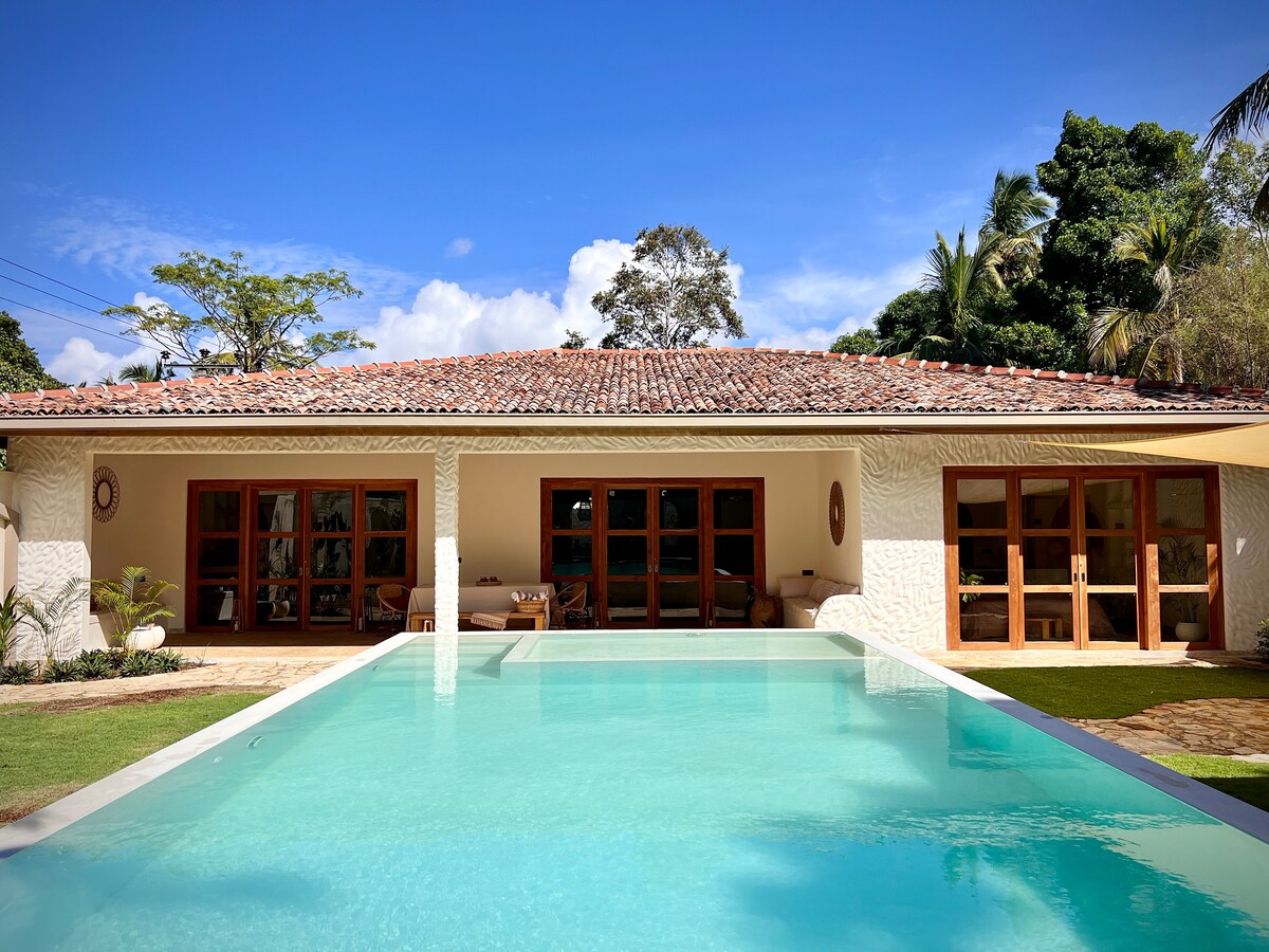A modern villa exterior is depicted, featuring large glass doors that provide a view of the inviting private pool. The pool reflects the clear blue sky, bordered by lush greenery and a well-maintained lawn. The traditional tiled roof adds character to the spacious structure.