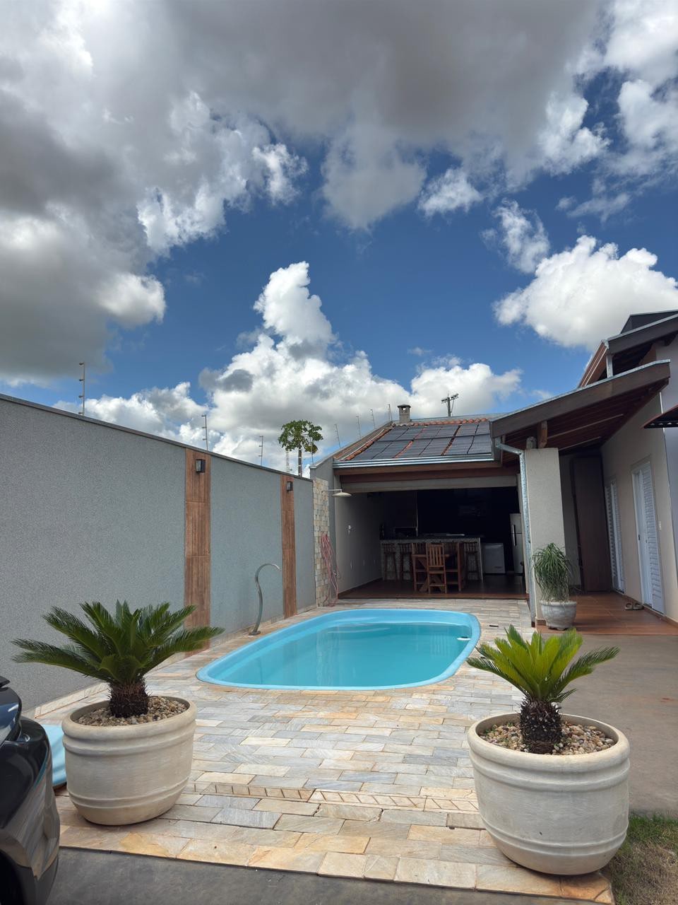 A swimming pool is situated in the foreground, surrounded by landscaped stone paths and potted plants. Behind the pool, an open space featuring large glass doors leads to a modern kitchen and dining area. A bright sky with scattered clouds complements the setting.