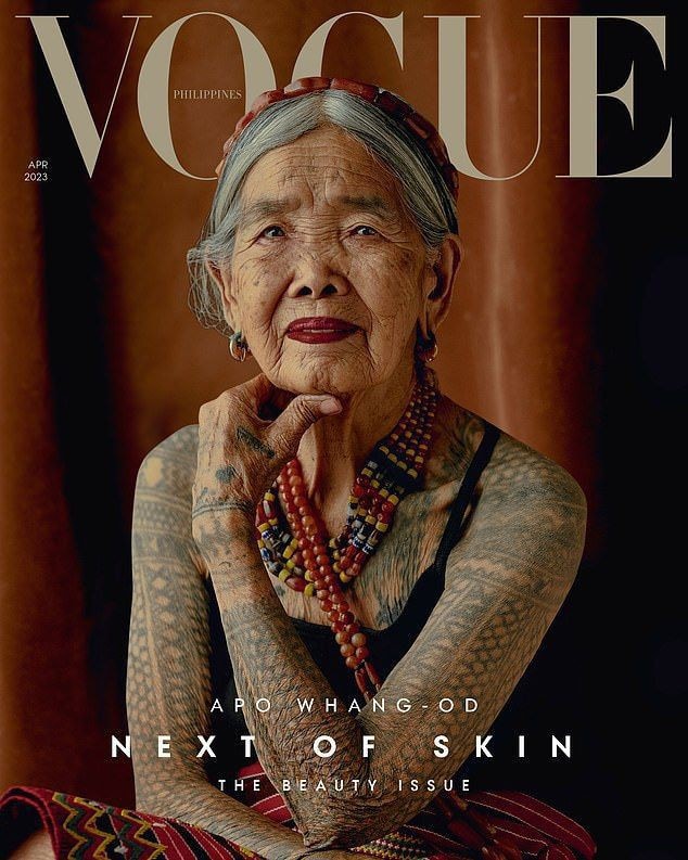 A woman with intricate tribal tattoos is seated against a warm, earthy backdrop. Her arms showcase detailed patterns, complemented by colorful beads adorning her neck. A serene expression reflects wisdom and tradition, highlighting the cultural significance of her artistry.