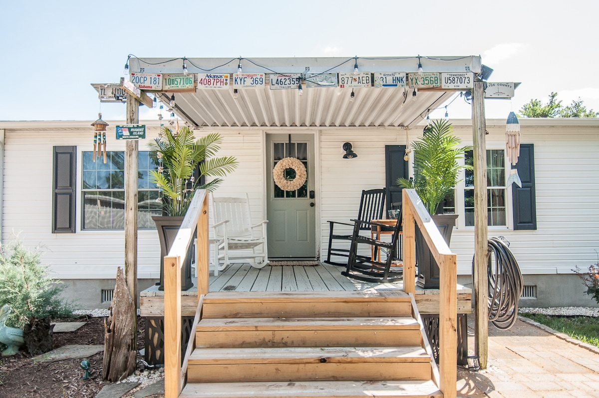 A welcoming entrance is featured, showcasing a spacious wooden deck with steps leading to a door. Rocking chairs are positioned under a covered area, surrounded by potted plants and decorative elements, including a wreath on the door and strings of lights overhead.