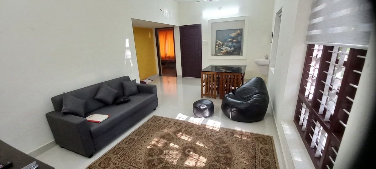 A bright living area features a comfortable gray sofa with decorative pillows, alongside a wooden table and a bean bag. Natural light filters through partially open blinds, illuminating the space. A patterned rug adds warmth to the polished floor.